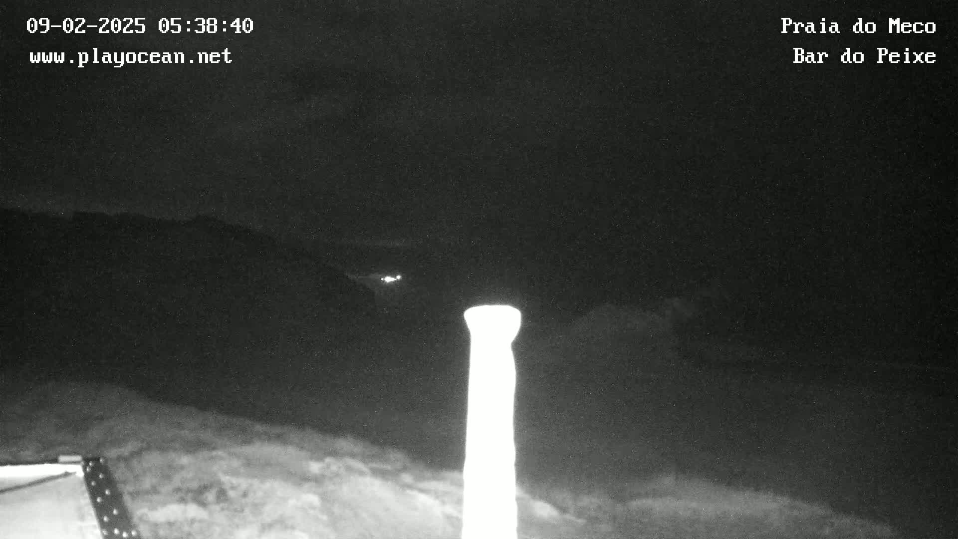 A nighttime, low-light image shows a tall, bright column-like object in the foreground against a dark, cloudy sky with faint distant lights.