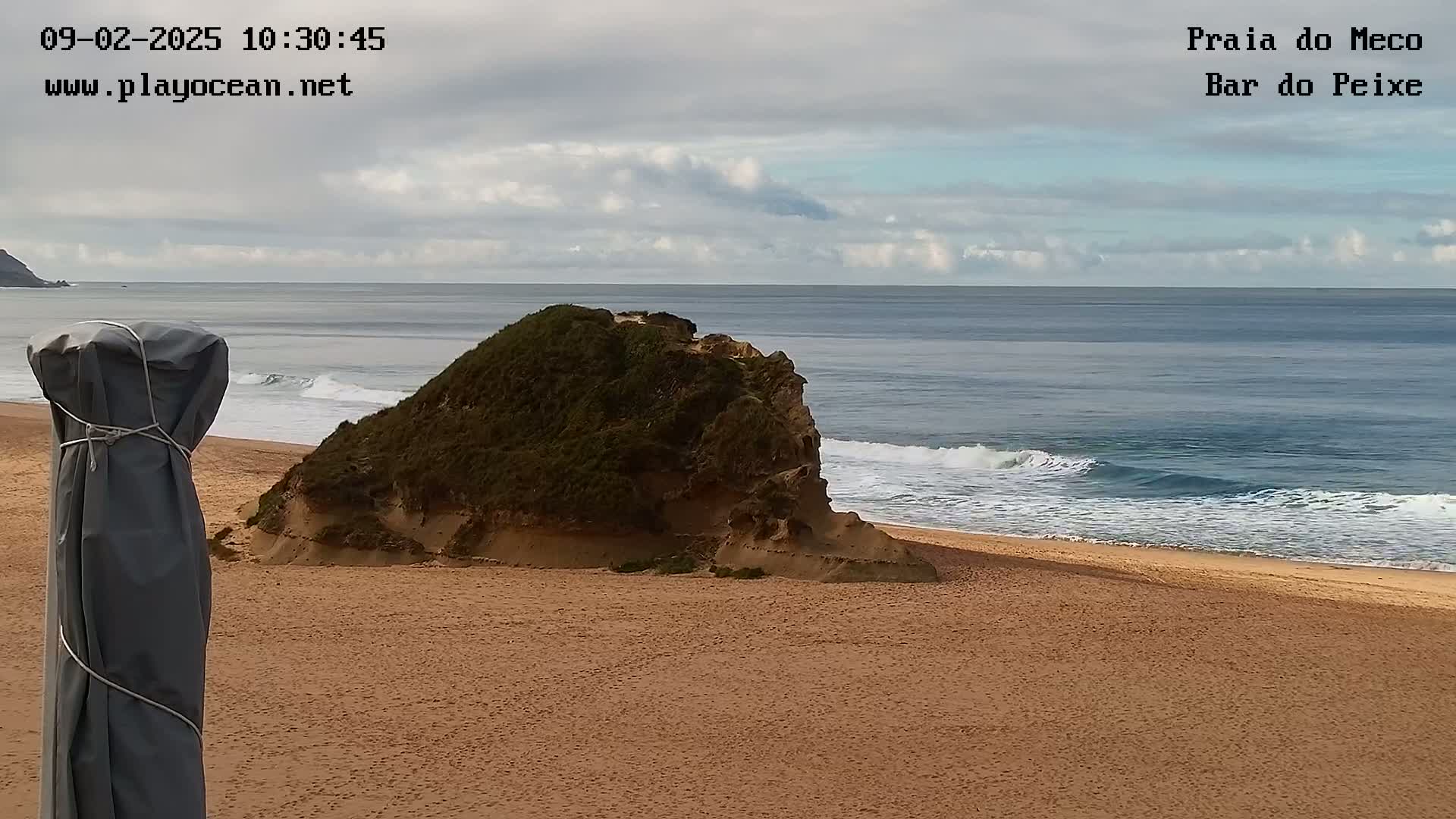 A sandy beach with a partially submerged rock formation sits under an overcast sky, with gentle waves rolling onto the shore.