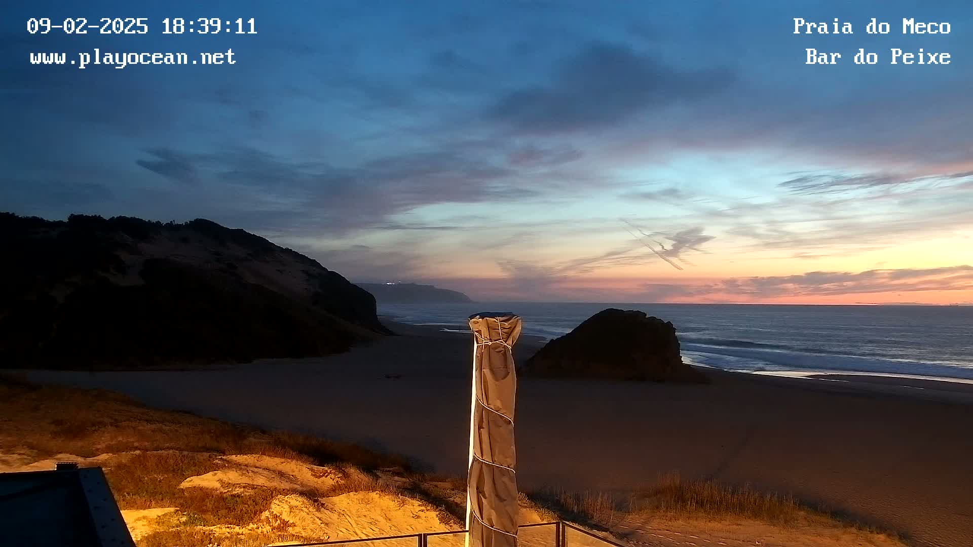 A sandy beach at sunset is seen from a dune, with a covered parasol in the foreground and dark cliffs and ocean in the background.