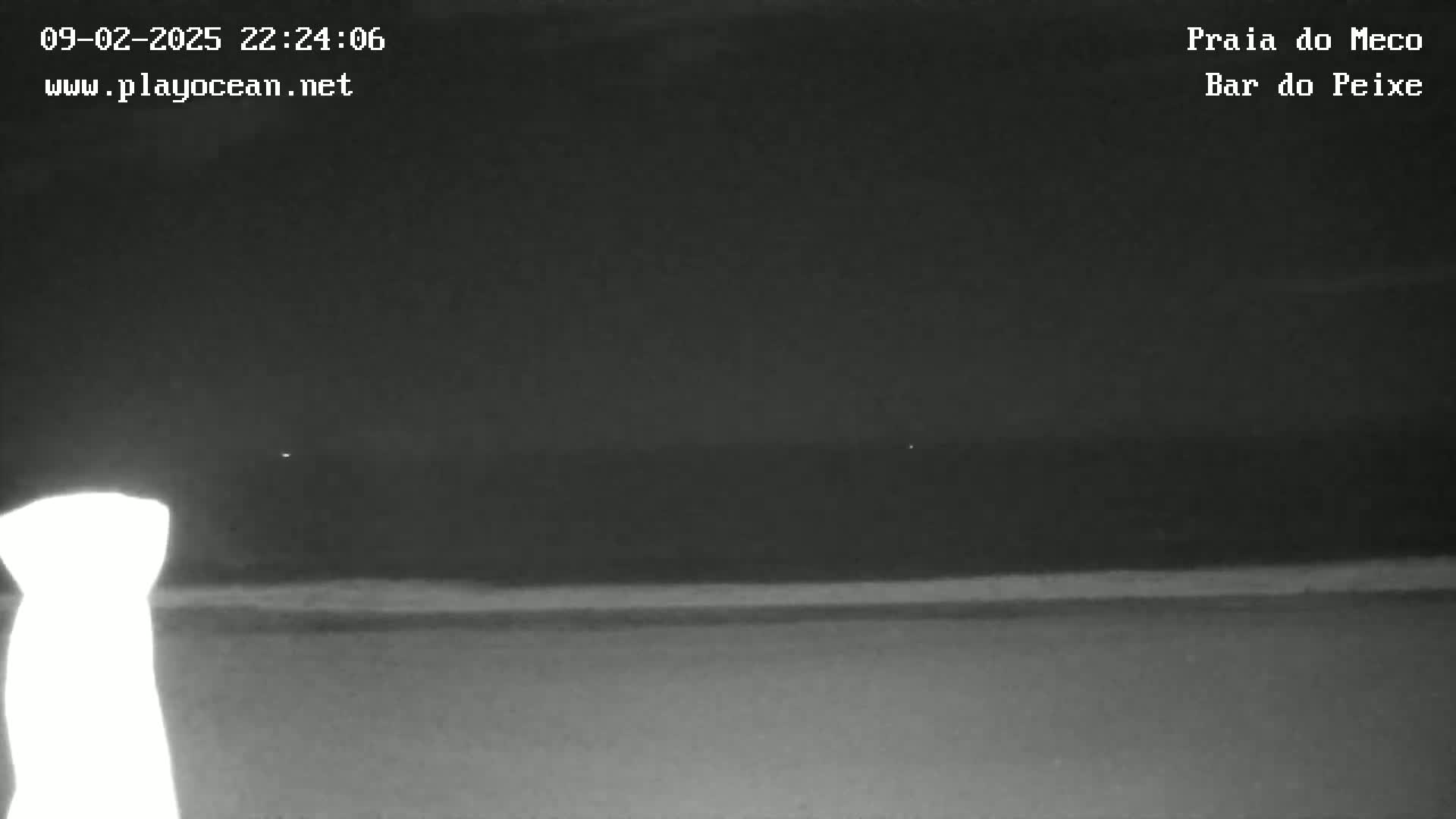 A nighttime, low-light view shows a dark ocean meeting a dark beach under a mostly clear sky.