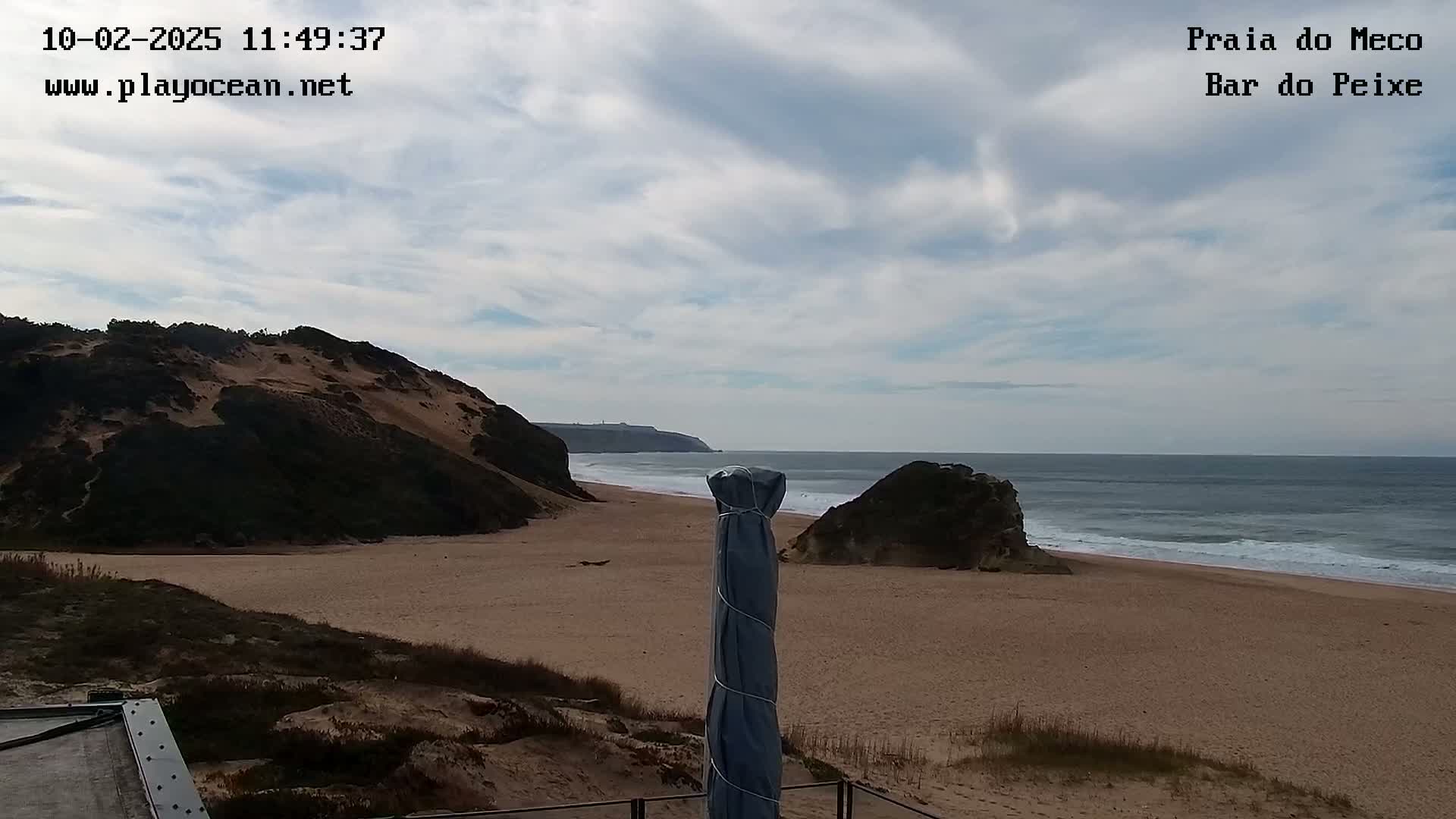 A sandy beach with a partly cloudy sky, backed by sand dunes and cliffs, stretches to a calm ocean with small waves.