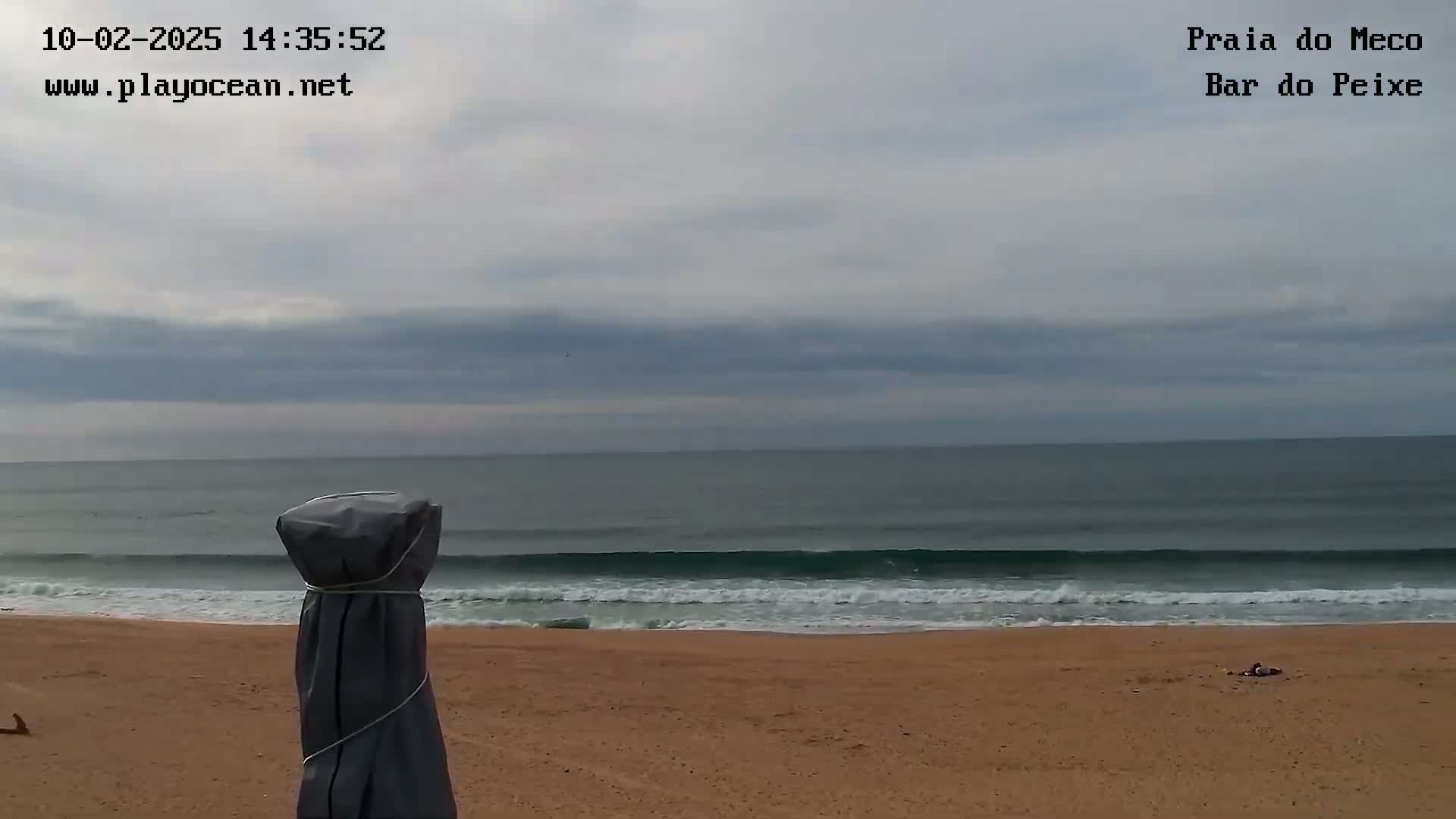 A sandy beach with a calm ocean under a mostly cloudy sky shows a covered beach umbrella in the foreground.