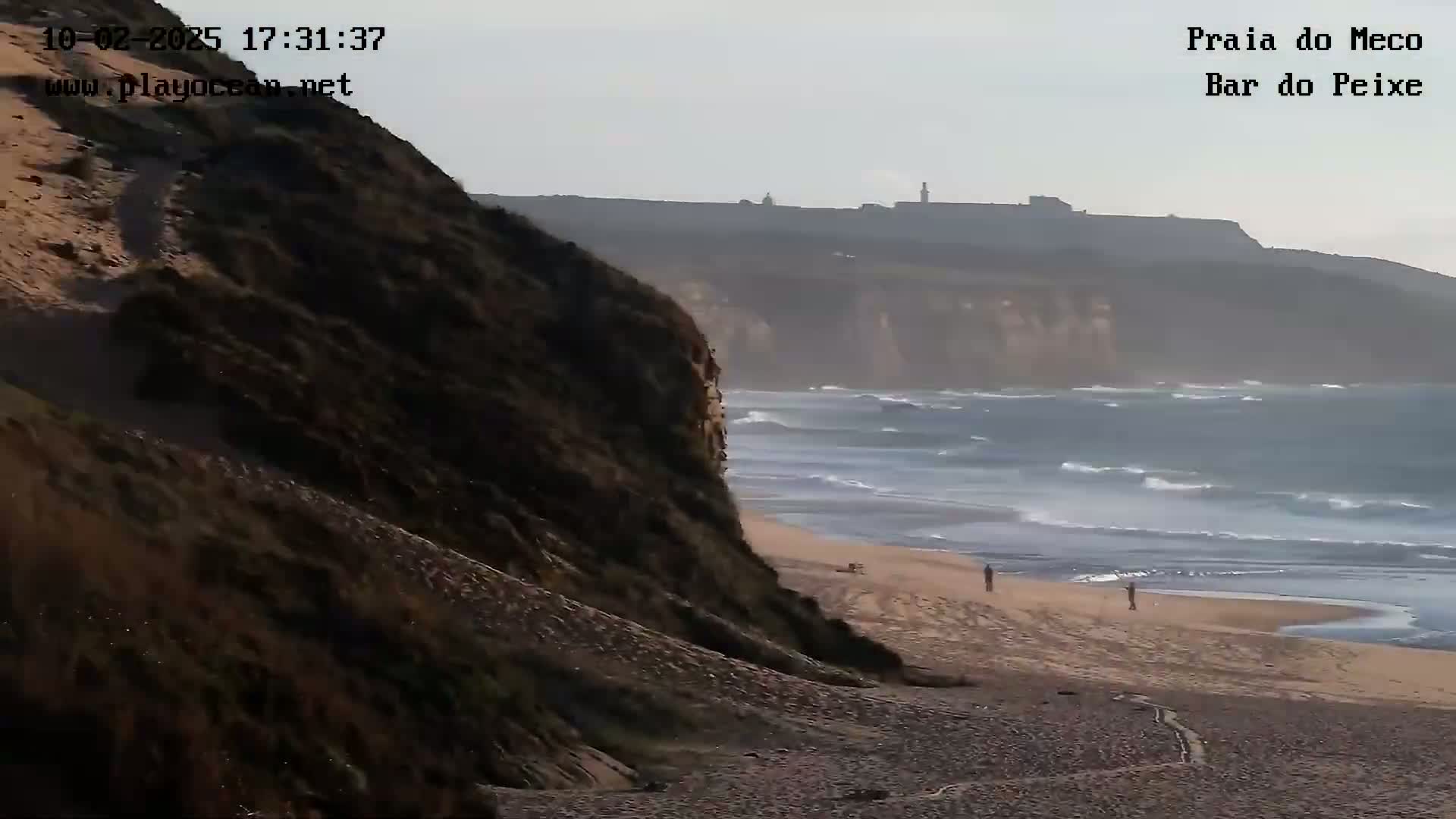 A sandy beach with a few people on it is situated below a steep cliff face, with somewhat choppy waves in the ocean and hazy conditions in the distance.