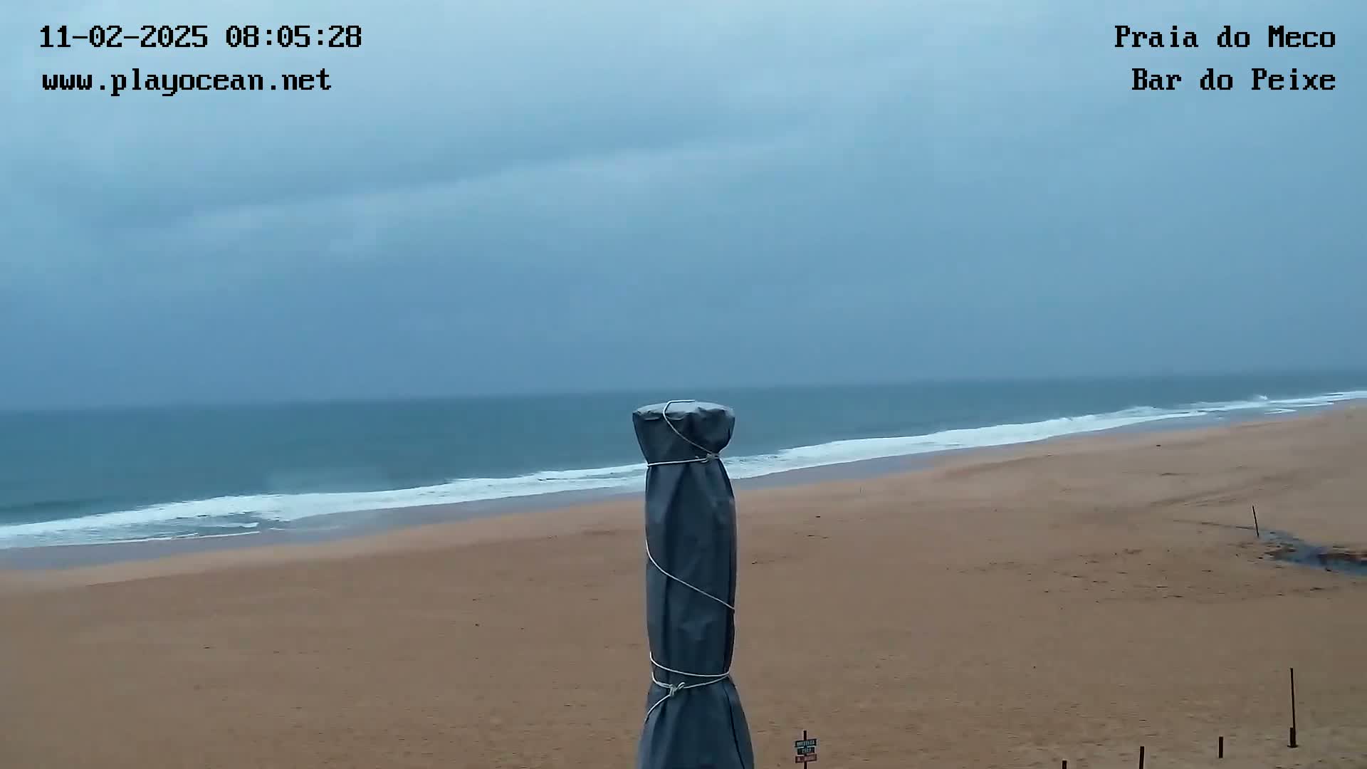 A sandy beach with gentle waves under an overcast sky is viewed from behind a covered beach umbrella.