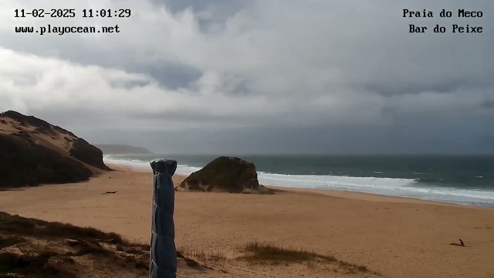 A sandy beach with moderate waves under a cloudy sky is seen from a dune, with a covered beach umbrella in the foreground and a rock formation visible near the water.