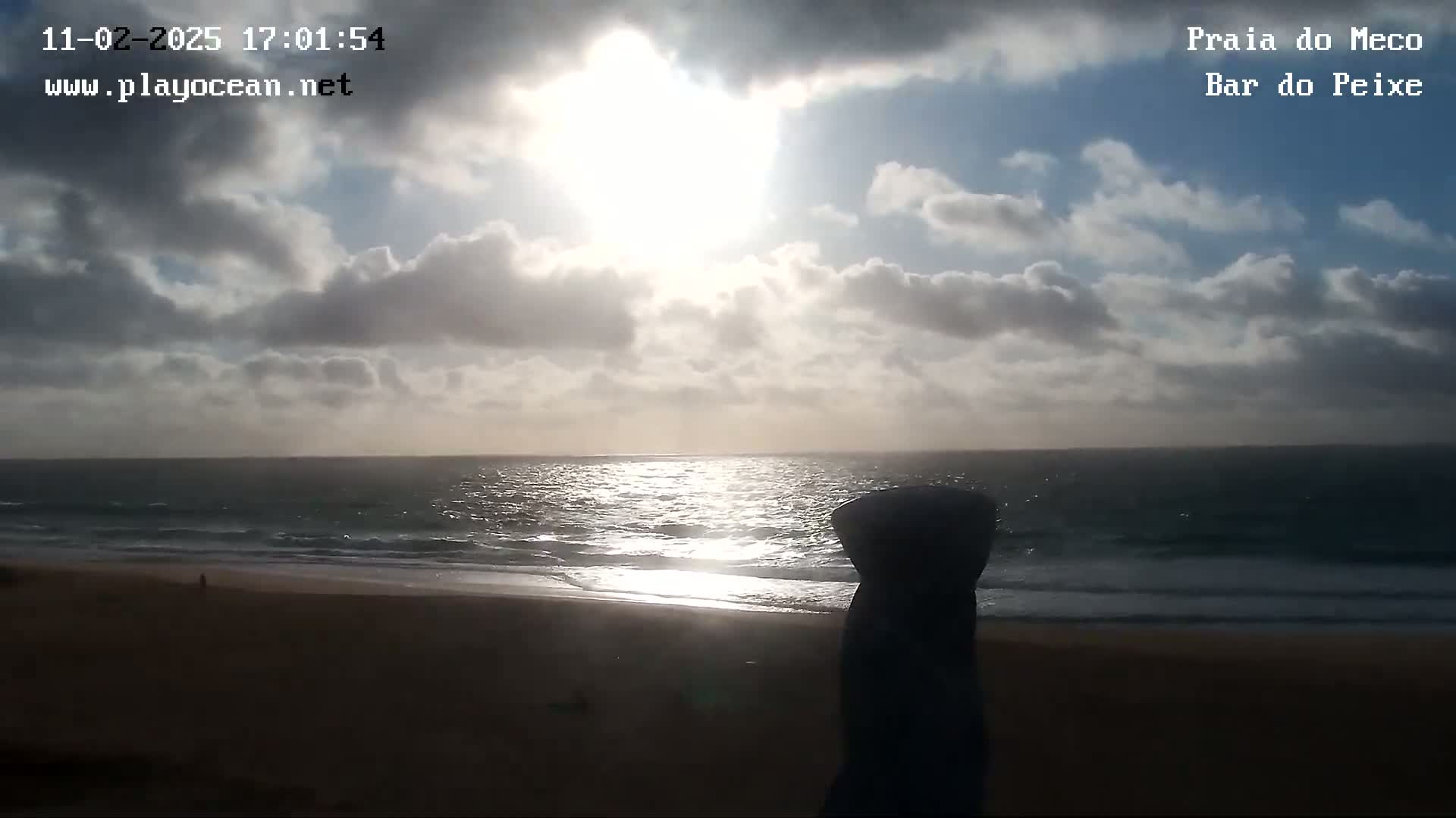 A partly cloudy day at a beach shows the ocean with some waves and a sandy shore, the sun shining brightly through the clouds.