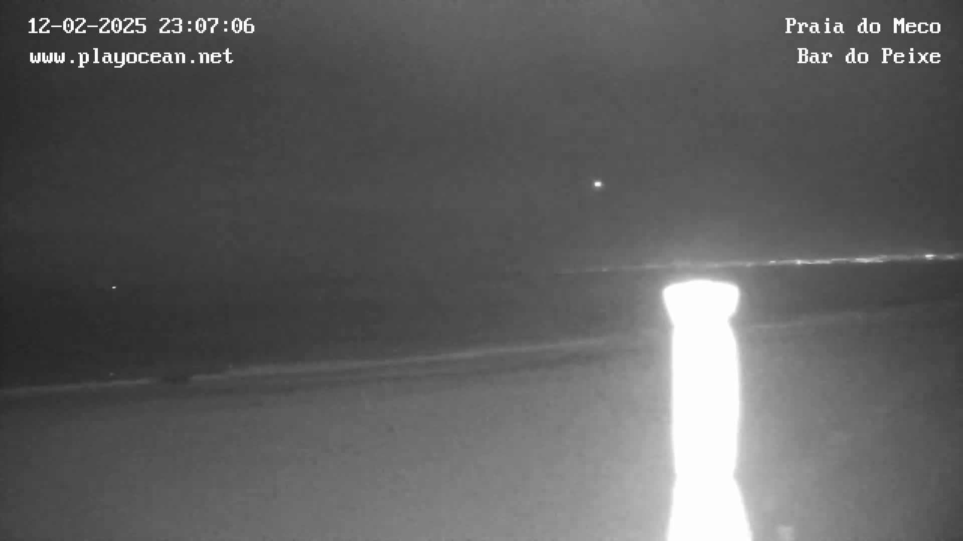 A nighttime, low-light view shows a dark beach and ocean with a bright light source in the foreground and a faint horizon line.