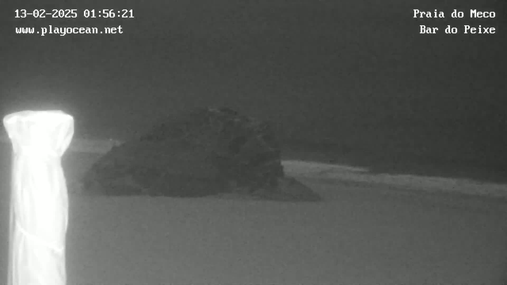A nighttime, low-light image shows a dark, rocky outcrop on a beach with a slightly visible wave line near the shore, and a bright, possibly reflective object in the foreground.