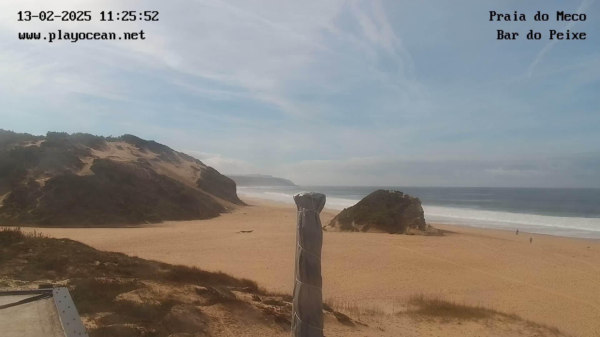 A sandy beach stretches between a dune-covered hill and the ocean under a mostly clear sky, with a few small waves breaking on the shore.