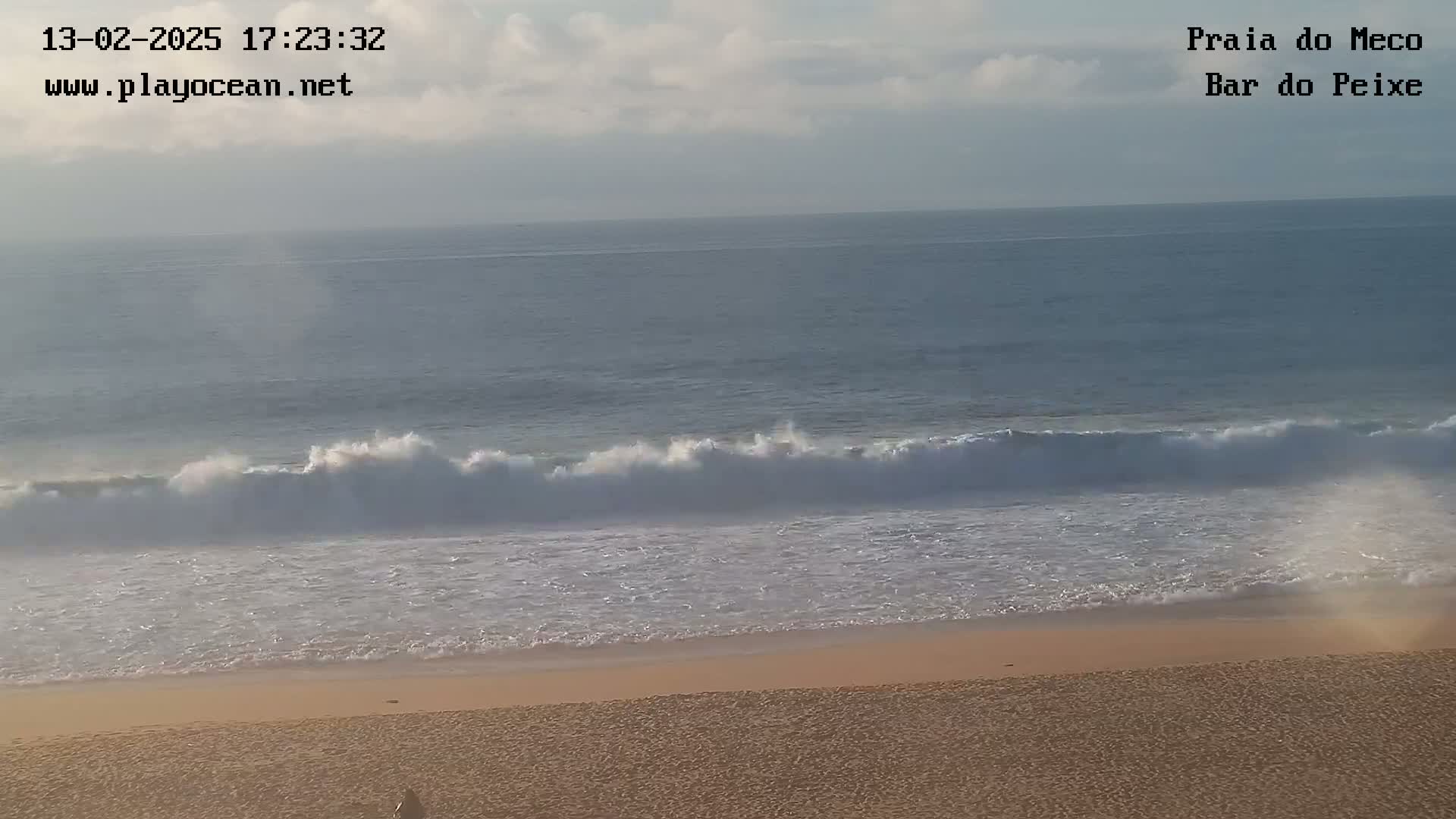 Ocean waves break on a sandy beach under a mostly cloudy sky.