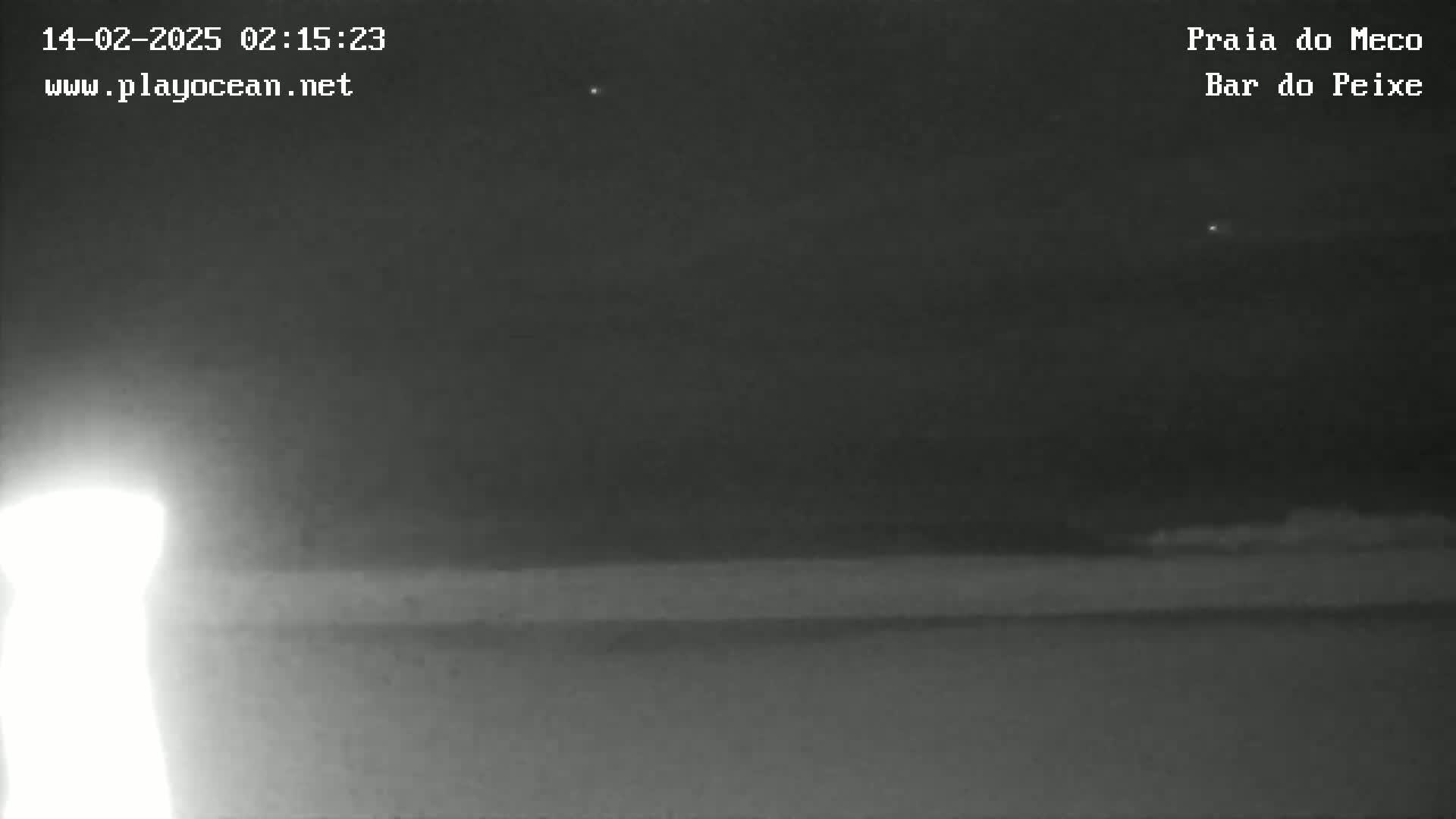 A nighttime, low-light image shows a dark beach and a bright light source in the lower left corner under a mostly clear sky.
