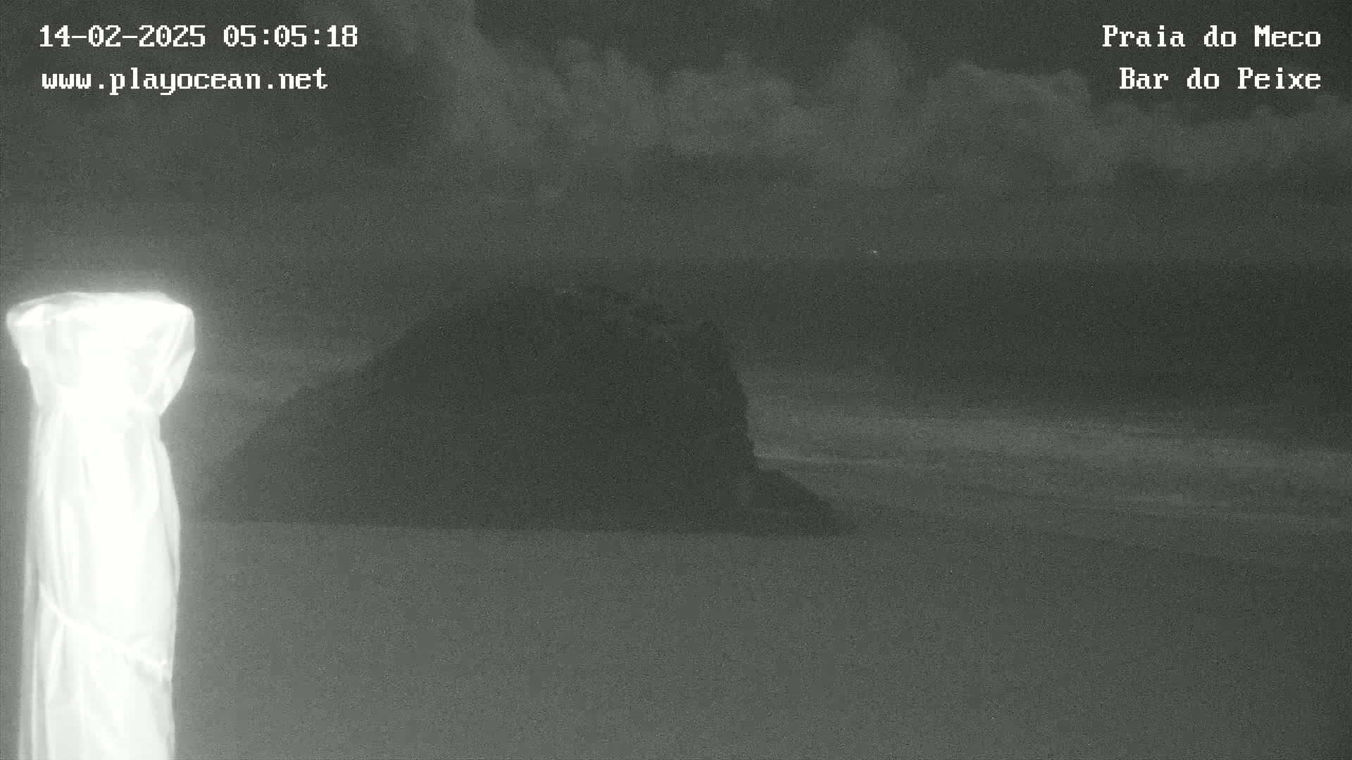 A dark, overcast night scene shows a large, dark rock formation near a shoreline with gently lapping waves.