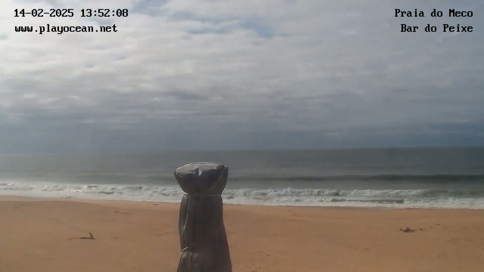 A mostly cloudy day shows a sandy beach with gentle waves rolling onto the shore, and a covered object in the foreground.