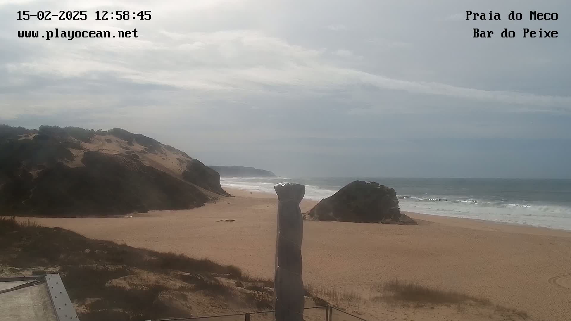 A sandy beach with a partly cloudy sky and gentle waves is seen from a vantage point overlooking a dune and a rock formation.