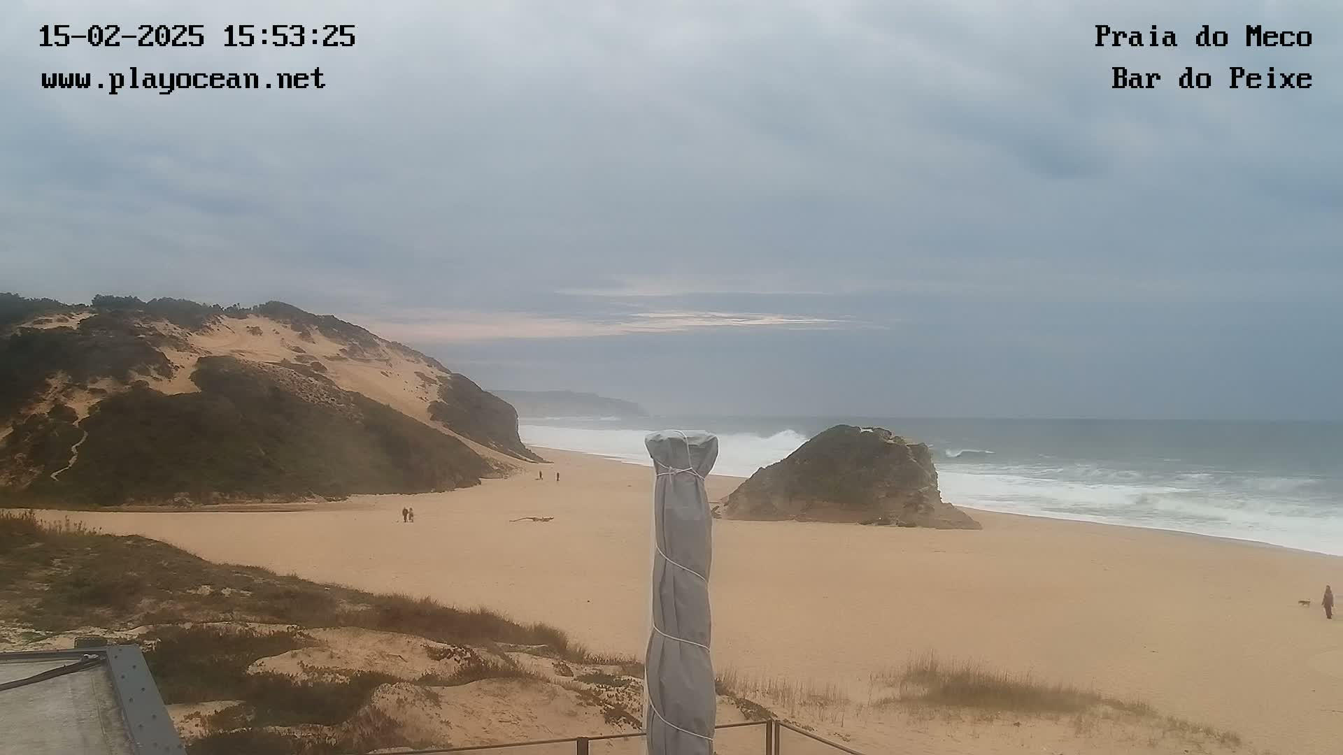A sandy beach with a large sand dune and rocky outcroppings is visible under an overcast sky, with moderate waves breaking along the shoreline.
