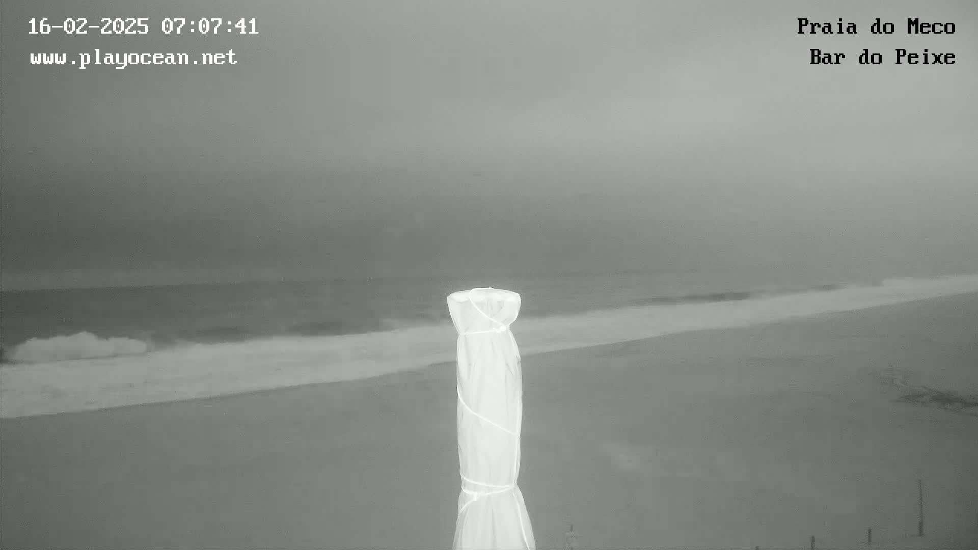 A grayscale image shows a beach with overcast skies and gently rolling waves, partially obscured by a covered beach umbrella in the foreground.