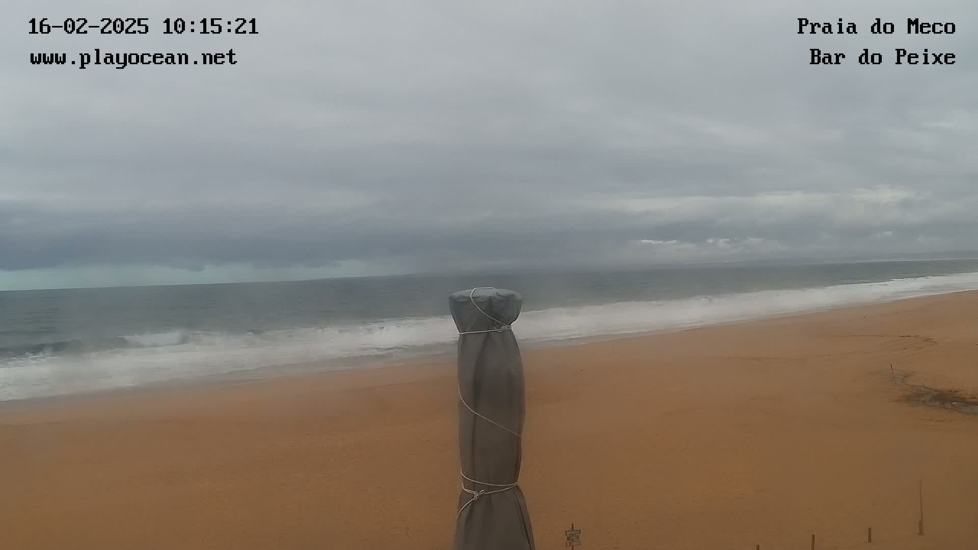A covered beach umbrella stands on a sandy beach under an overcast sky, with waves rolling in from a gray ocean.
