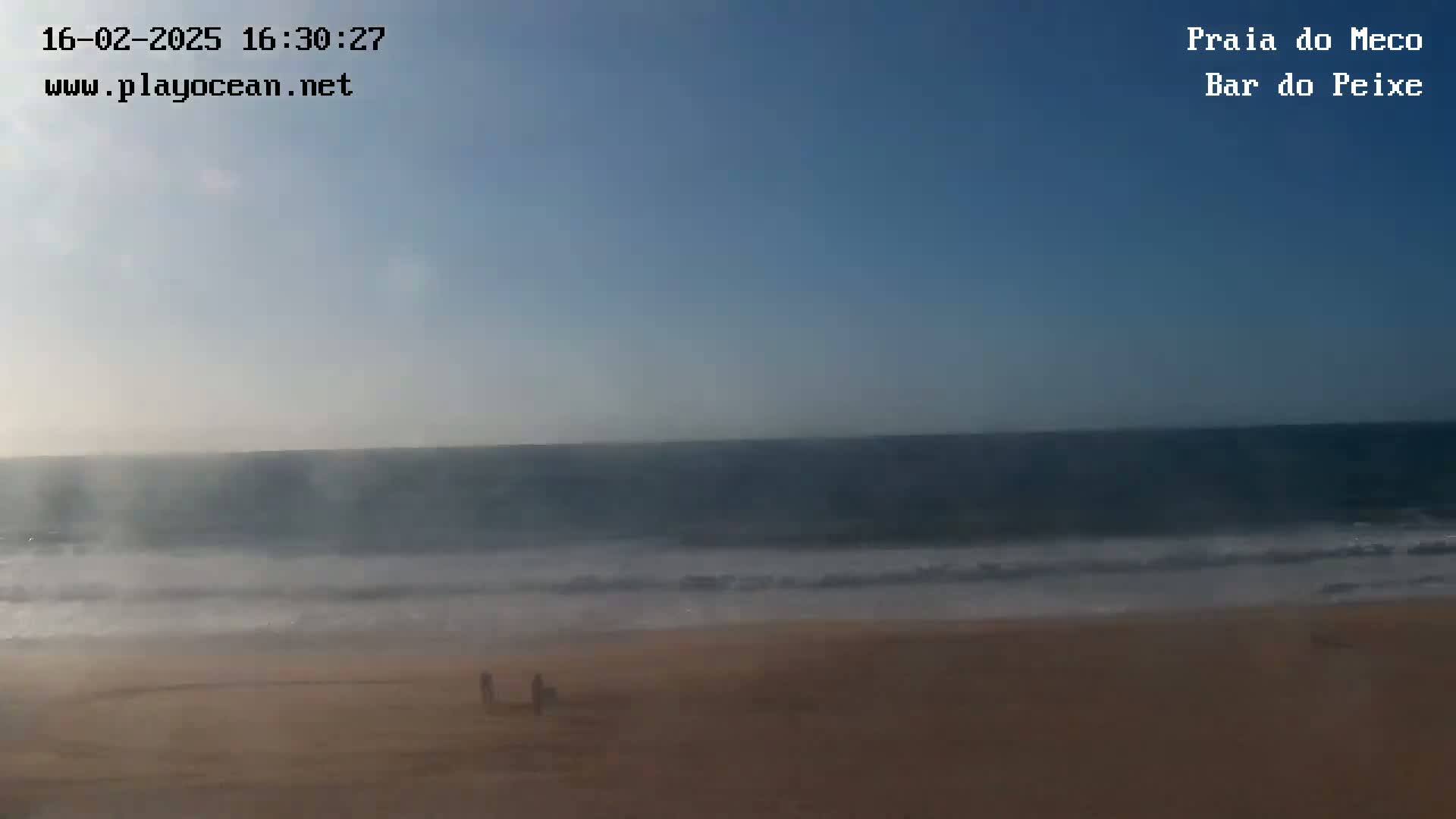A hazy, sunny day shows a calm ocean meeting a sandy beach where a few people are visible.