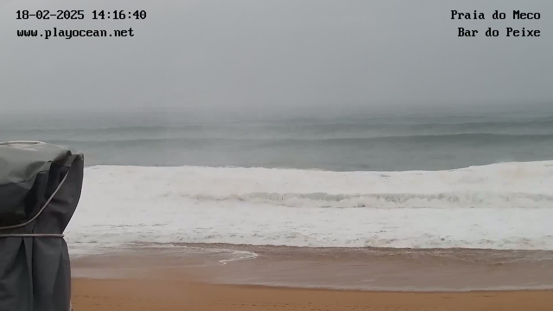 A sandy beach is shown with large, white-capped waves crashing on the shore under an overcast, gray sky.