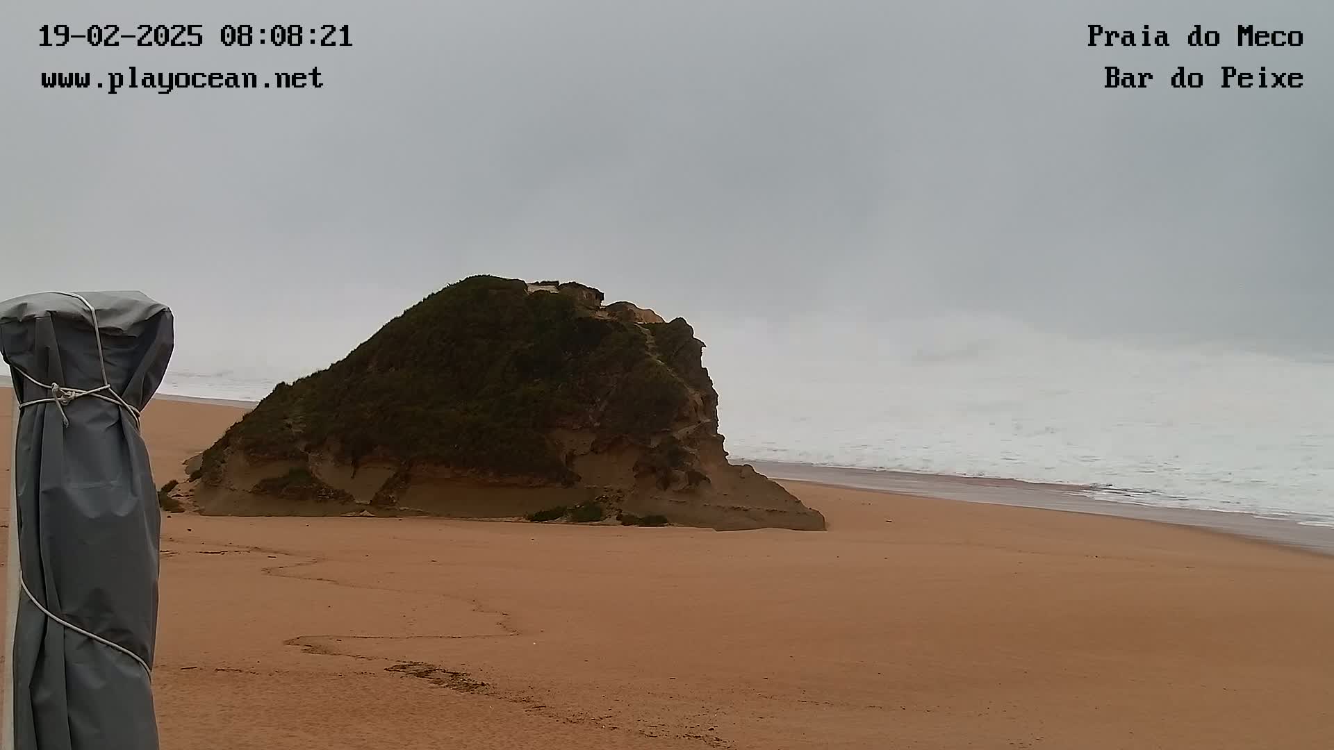 A sandy beach with a rocky outcrop is shown under an overcast sky, with moderate waves rolling onto the shore.