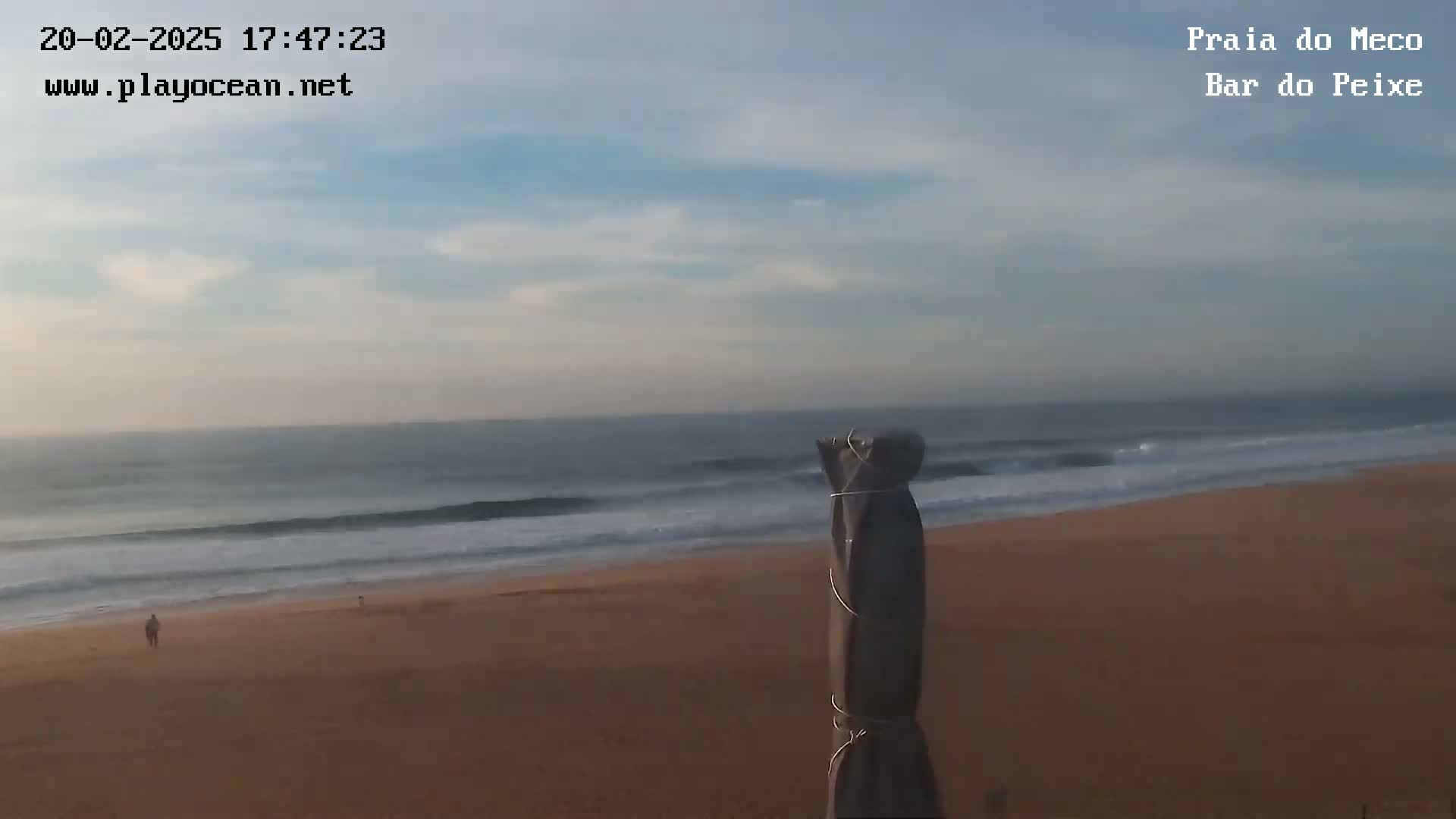 A mostly cloudy sky overlooks a sandy beach with gently rolling waves and a single person walking along the shore.