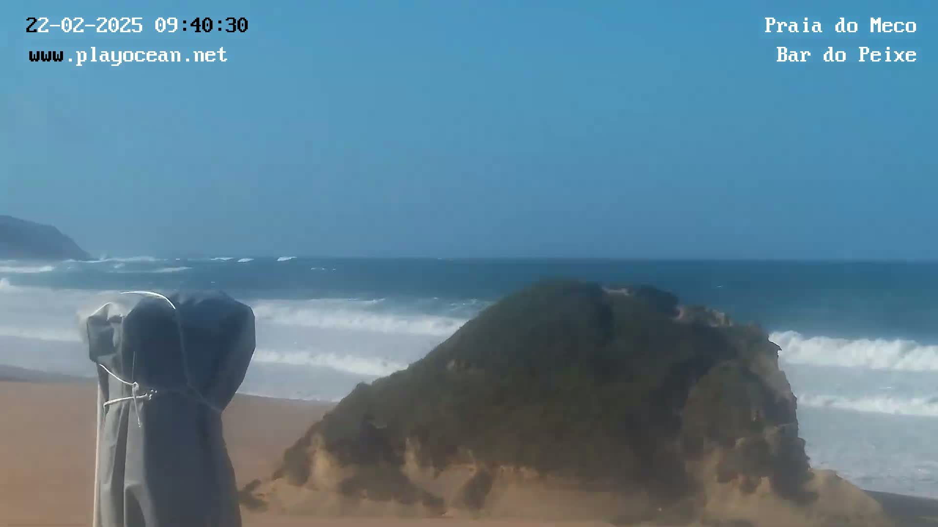 A sandy beach with a covered beach umbrella is shown, with a rocky outcropping in the foreground and a somewhat choppy ocean under a clear blue sky.