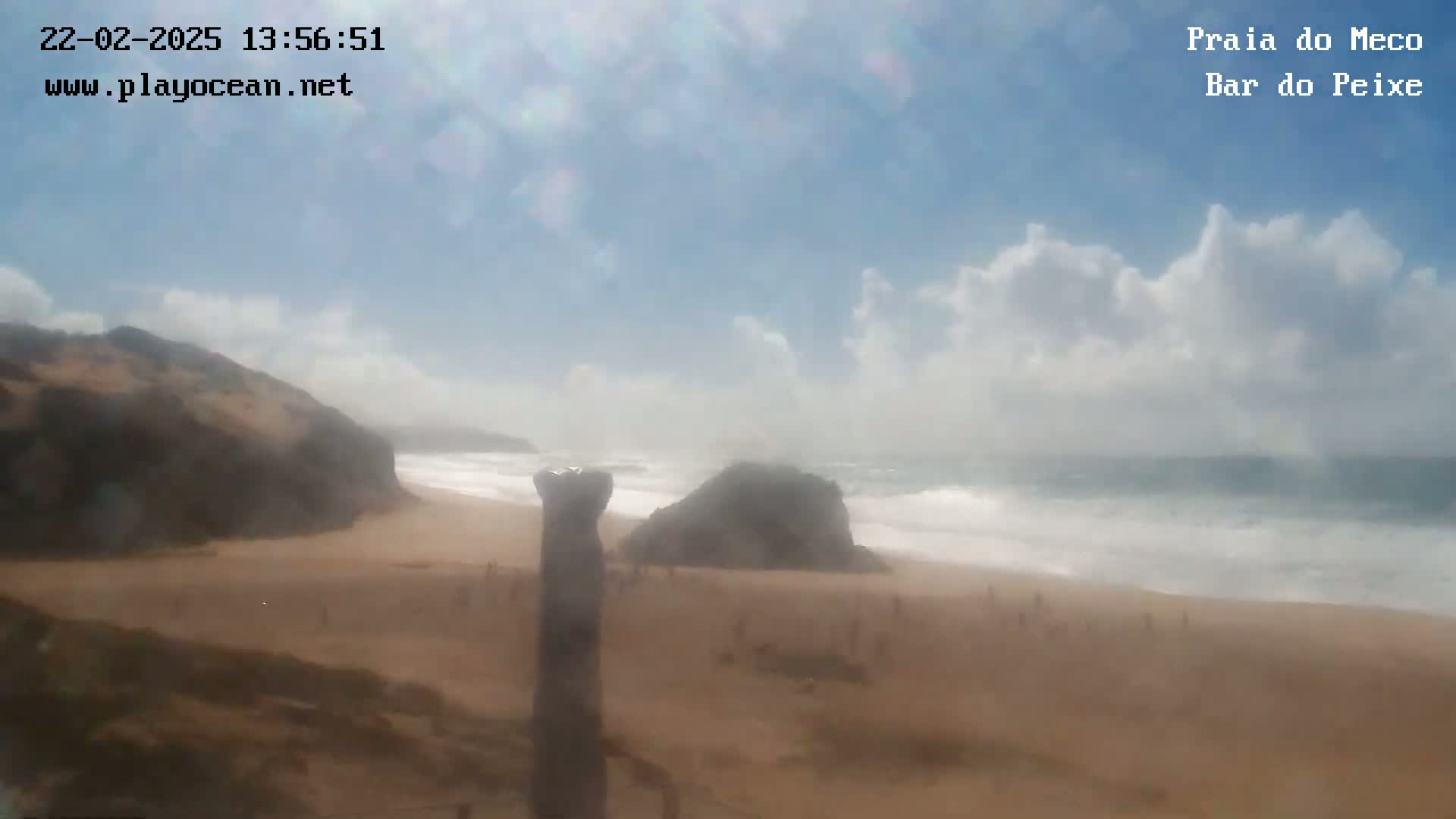 A sandy beach with a few people on it is partially obscured by hazy weather conditions, with waves breaking on the shore under a partly cloudy sky.