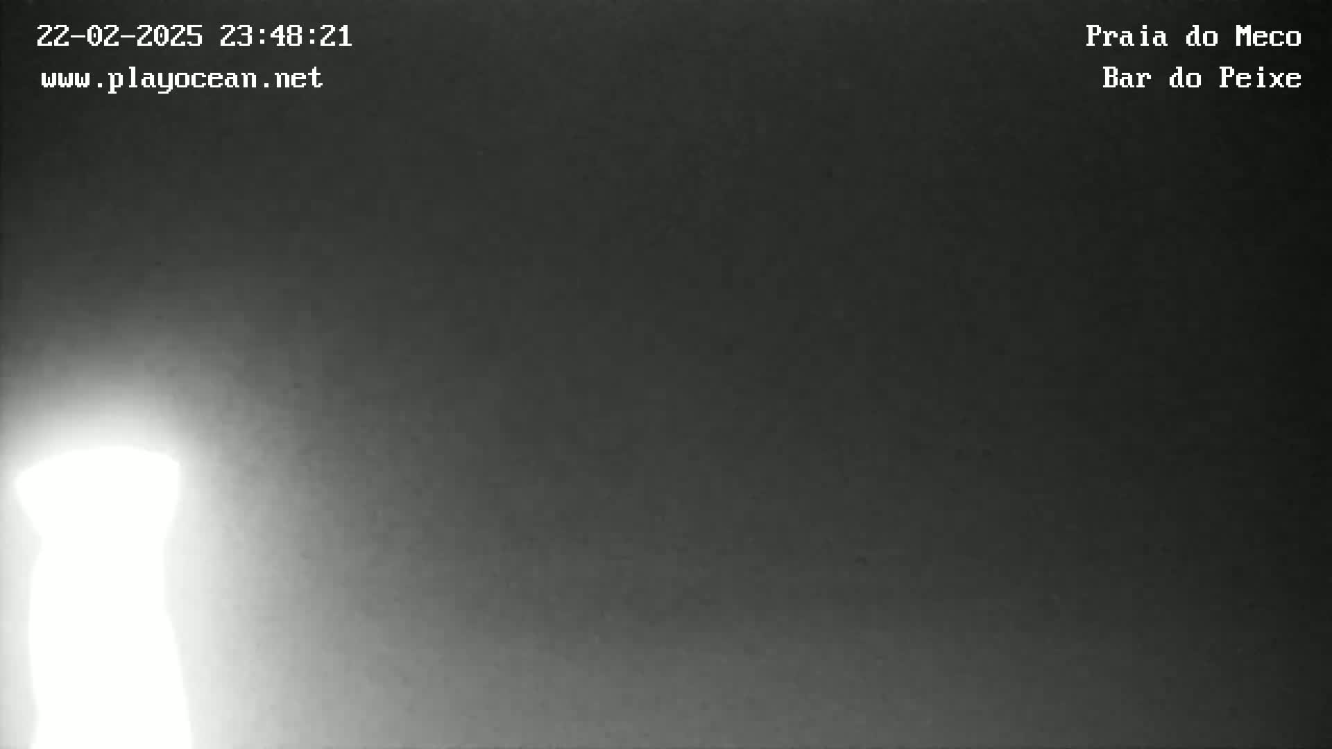 A bright light source, possibly a lamppost or lighthouse, is visible in the lower left corner of a dark, nighttime scene.