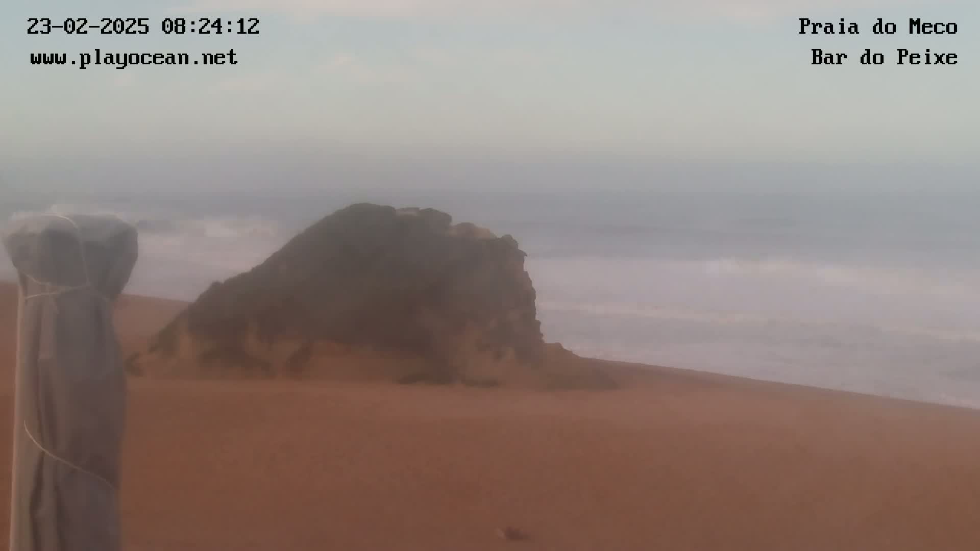 A hazy view shows a rocky outcrop on a sandy beach with small waves breaking in the ocean.