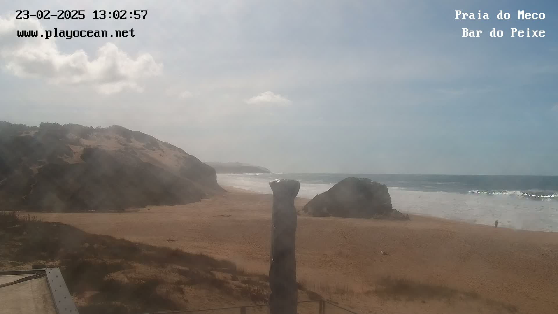 A sandy beach with rocky cliffs and a partly cloudy sky stretches to a gently wavy ocean.