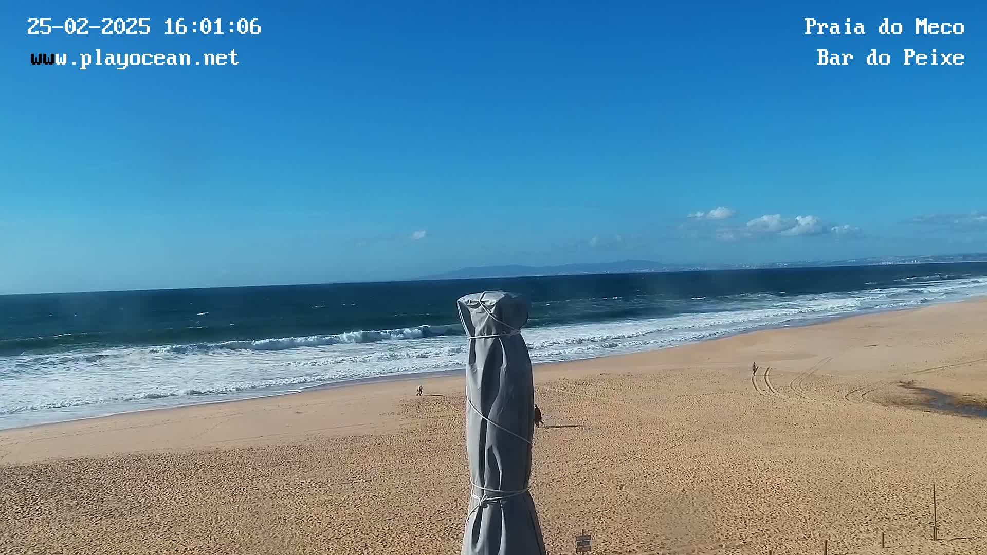 A sandy beach with a few people walking on it, moderate waves breaking on the shore under a clear blue sky, and a covered beach umbrella in the foreground.