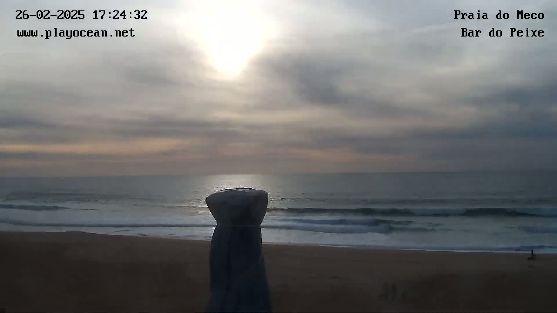 A mostly cloudy sunset over a calm ocean beach is viewed from behind a partially covered object.