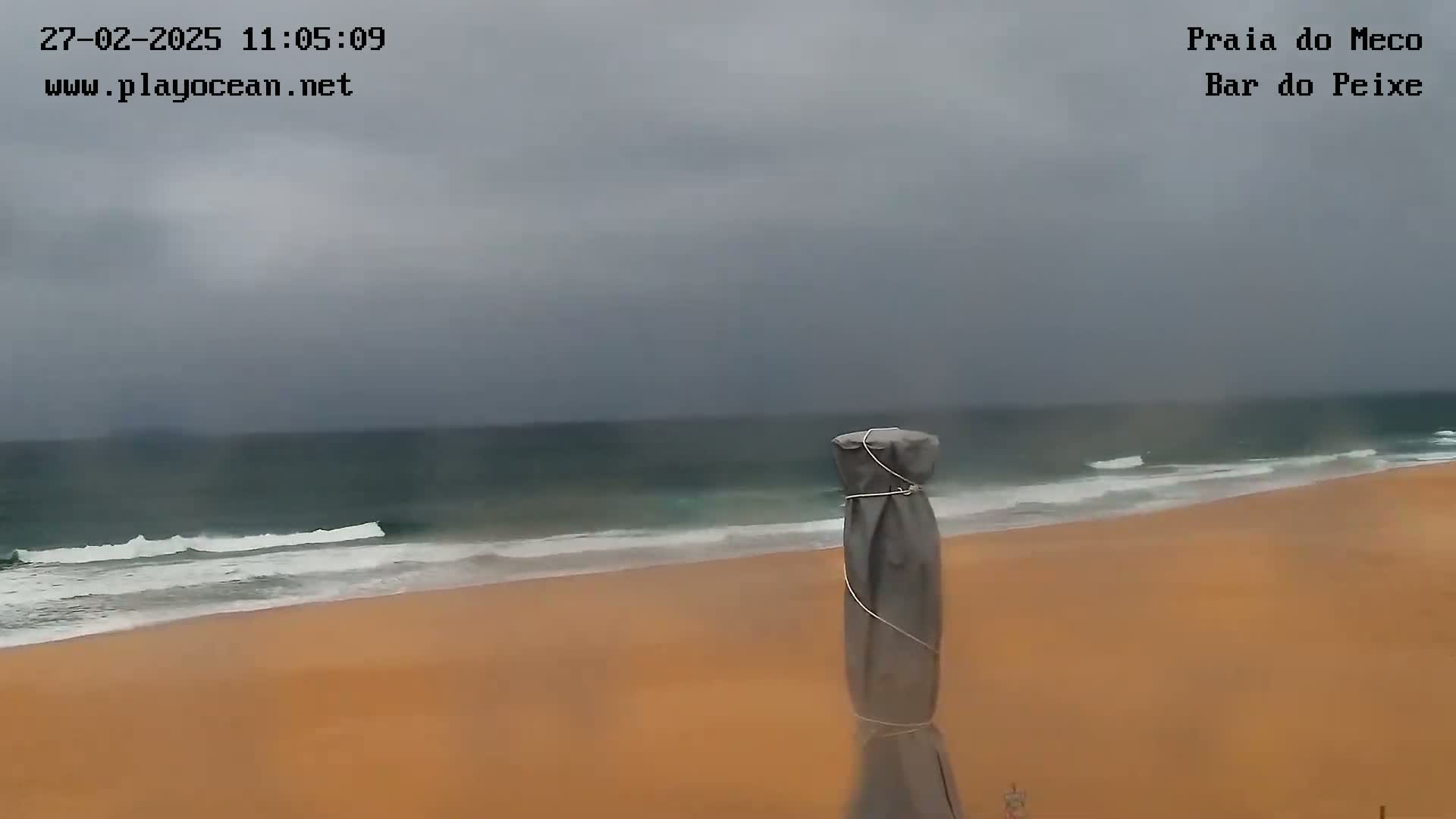 A sandy beach with moderate waves under an overcast sky is seen from a slightly elevated vantage point, near a covered object that looks like a beach umbrella.