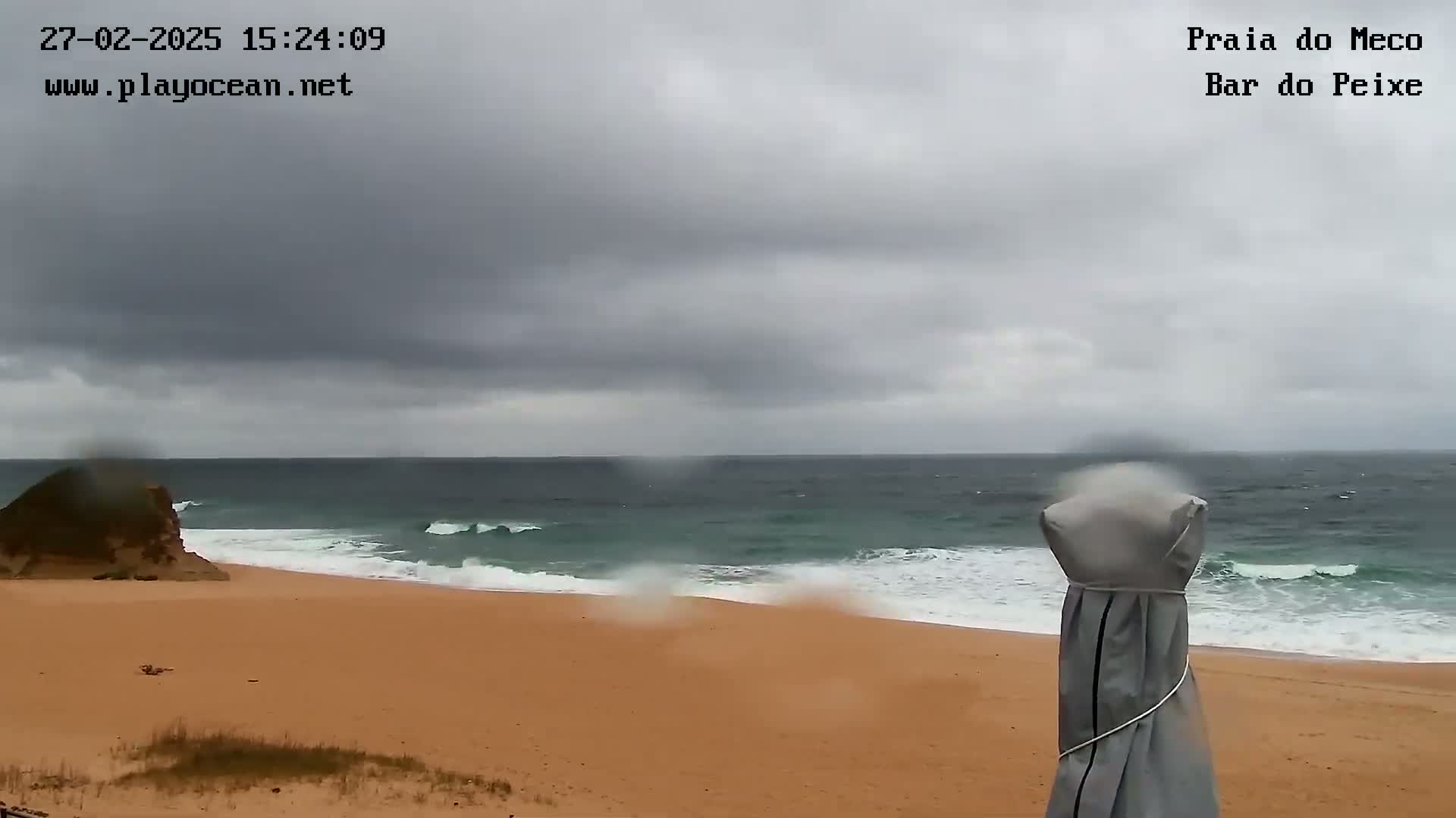 A sandy beach with moderate waves under an overcast sky is seen from a slightly elevated viewpoint, with rain visible on the camera lens and a covered beach umbrella in the foreground.