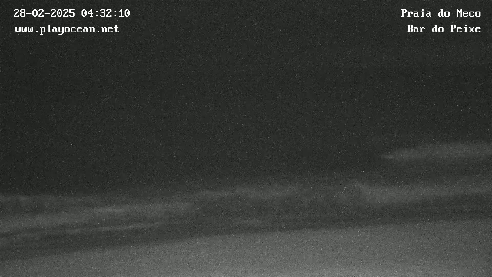 A dark, nighttime beach scene shows a mostly flat, dark expanse of sand and water with faint, blurry waves under a dark sky.