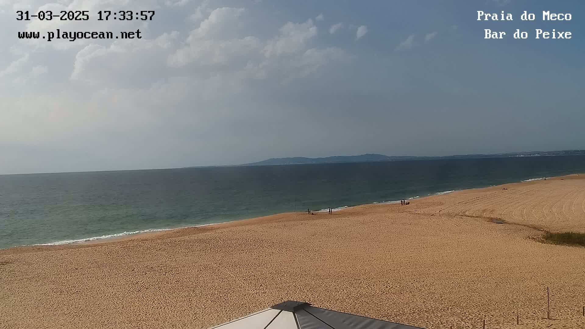A mostly sunny day at a sandy beach shows a calm ocean with a distant landmass under a partly cloudy sky.