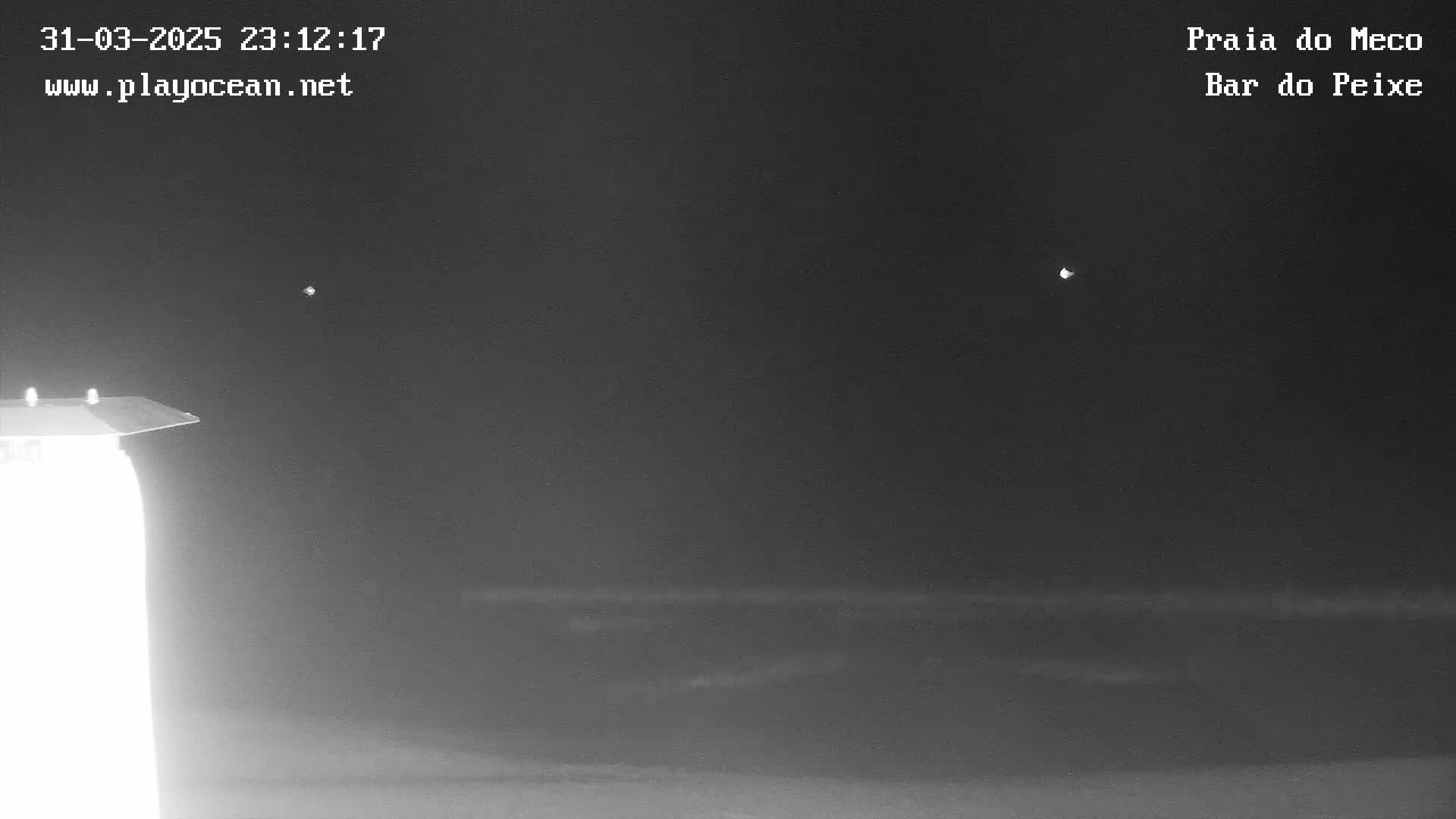 A nighttime, low-light view shows a dark ocean and a partially illuminated structure in the lower left corner under a clear, dark sky with two distant, bright lights.