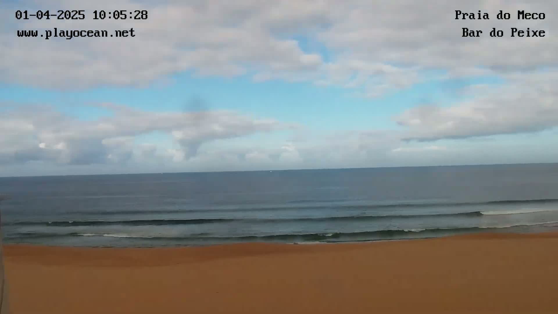 A mostly cloudy sky overlooks a calm ocean with small waves lapping a sandy beach.