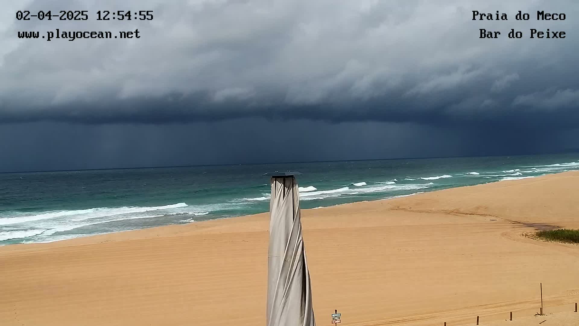 A sandy beach with a teal ocean under a dark, stormy sky features a tall, spiraling beach umbrella in the foreground.