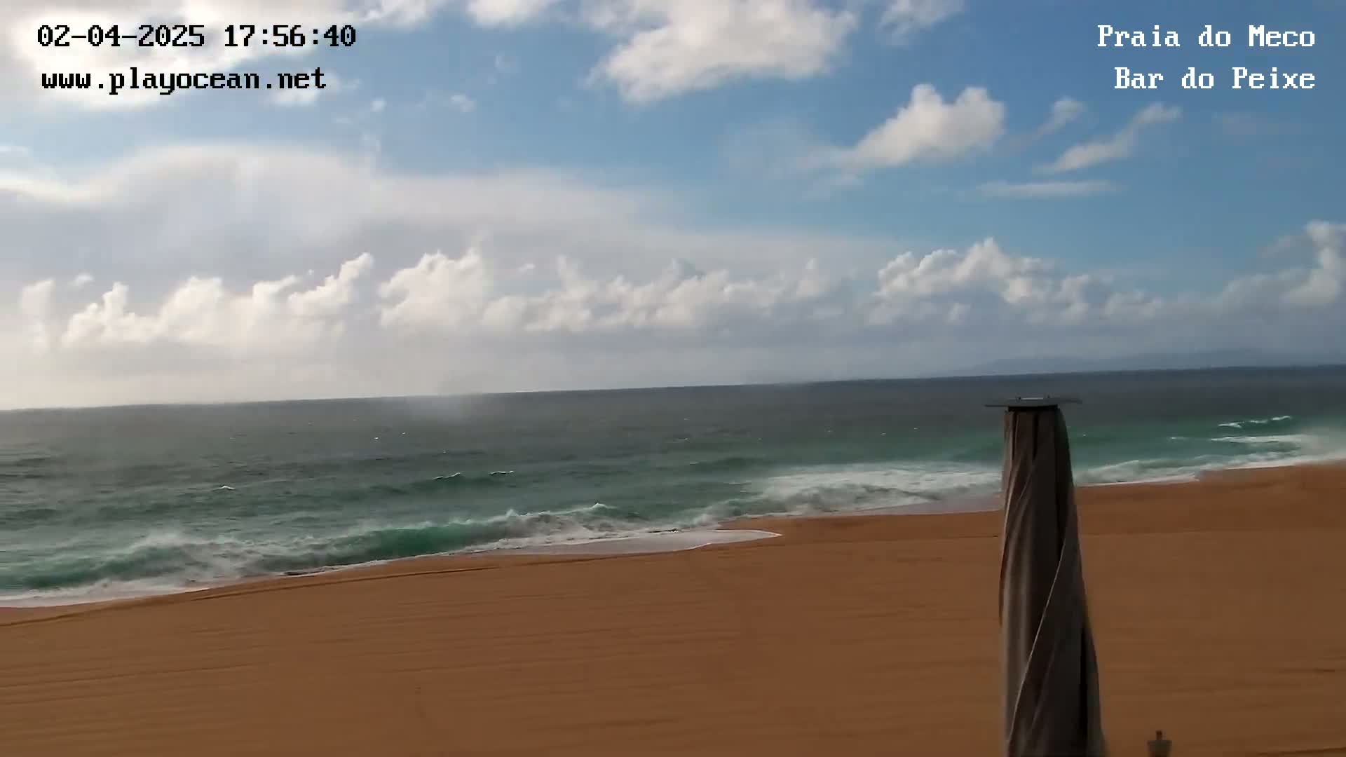 A partly cloudy sky overlooks a sandy beach with moderate waves rolling onto the shore.