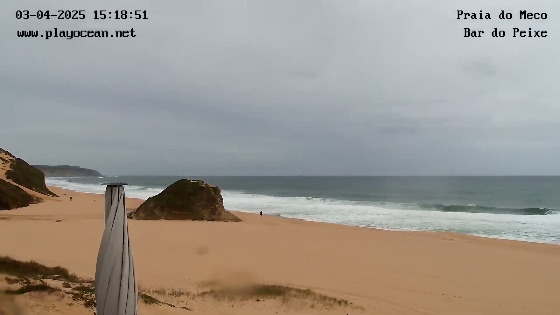 A sandy beach with a few people, a rocky outcrop, and gentle waves under an overcast sky.