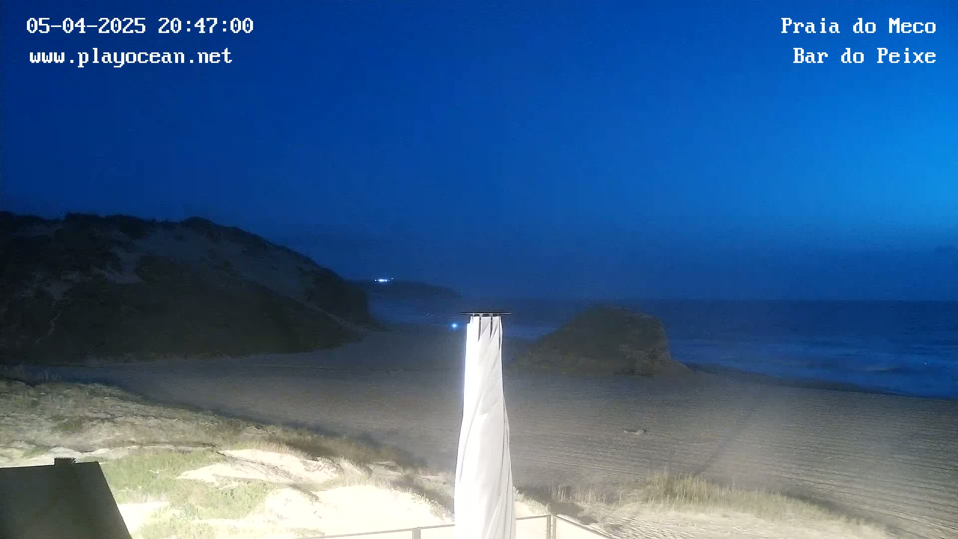 A sandy beach at twilight under a clear, dark blue sky, with a tall, white beach umbrella in the foreground.