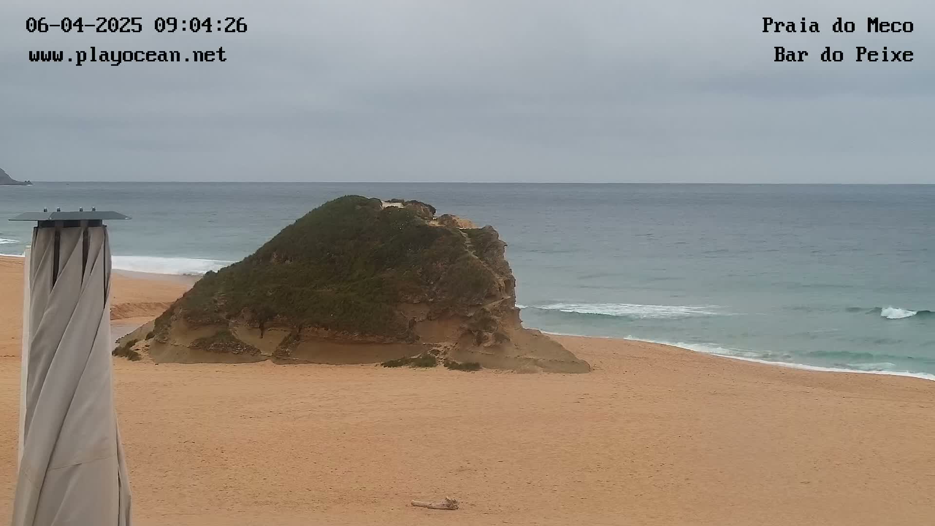 A sandy beach with a small, green-topped rock formation sits near the water's edge under an overcast sky.