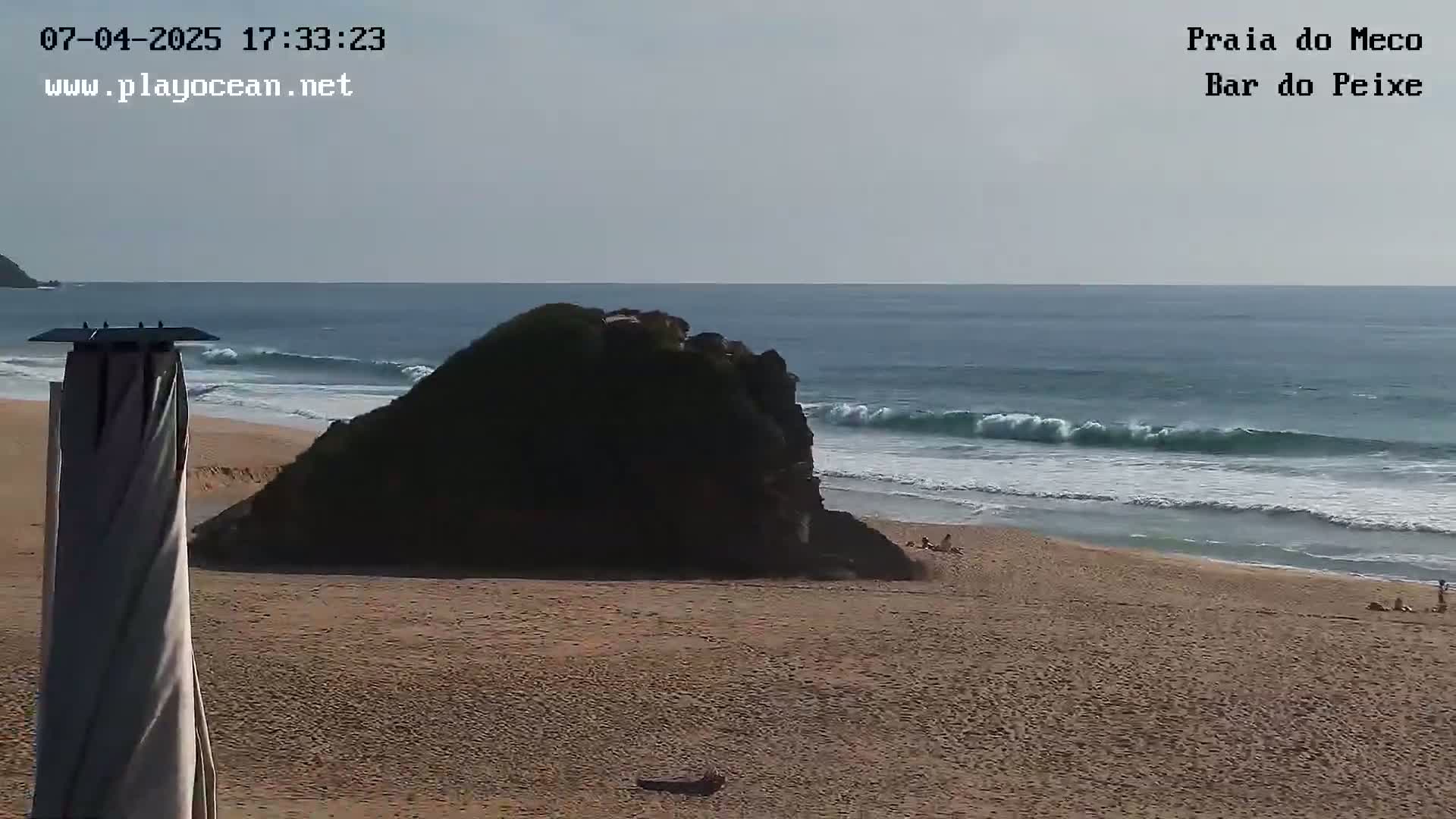 A sandy beach with a large dark rock formation near the water’s edge, gentle waves rolling onto the shore under a mostly clear sky.
