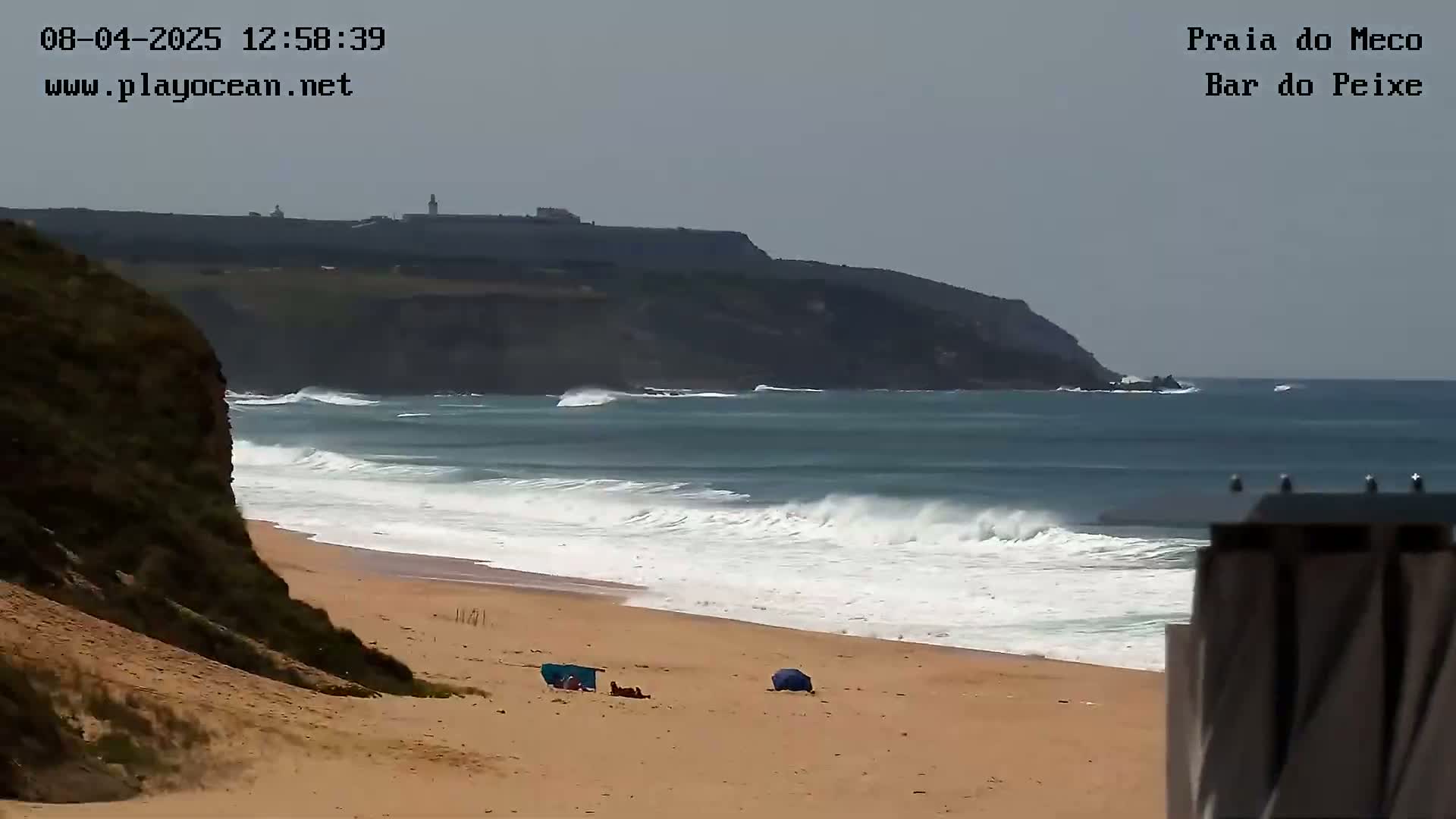 A sandy beach with a few people and beach umbrellas is shown, with moderate waves rolling in from an ocean under a mostly clear sky, and a distant cliff visible.