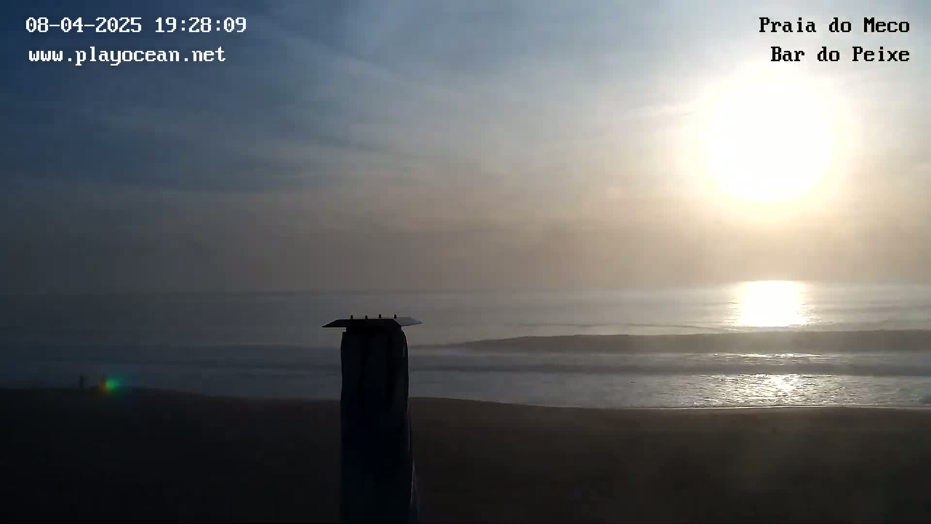 A hazy sunset over a dark beach with gentle waves, viewed from behind a tall, slender structure.