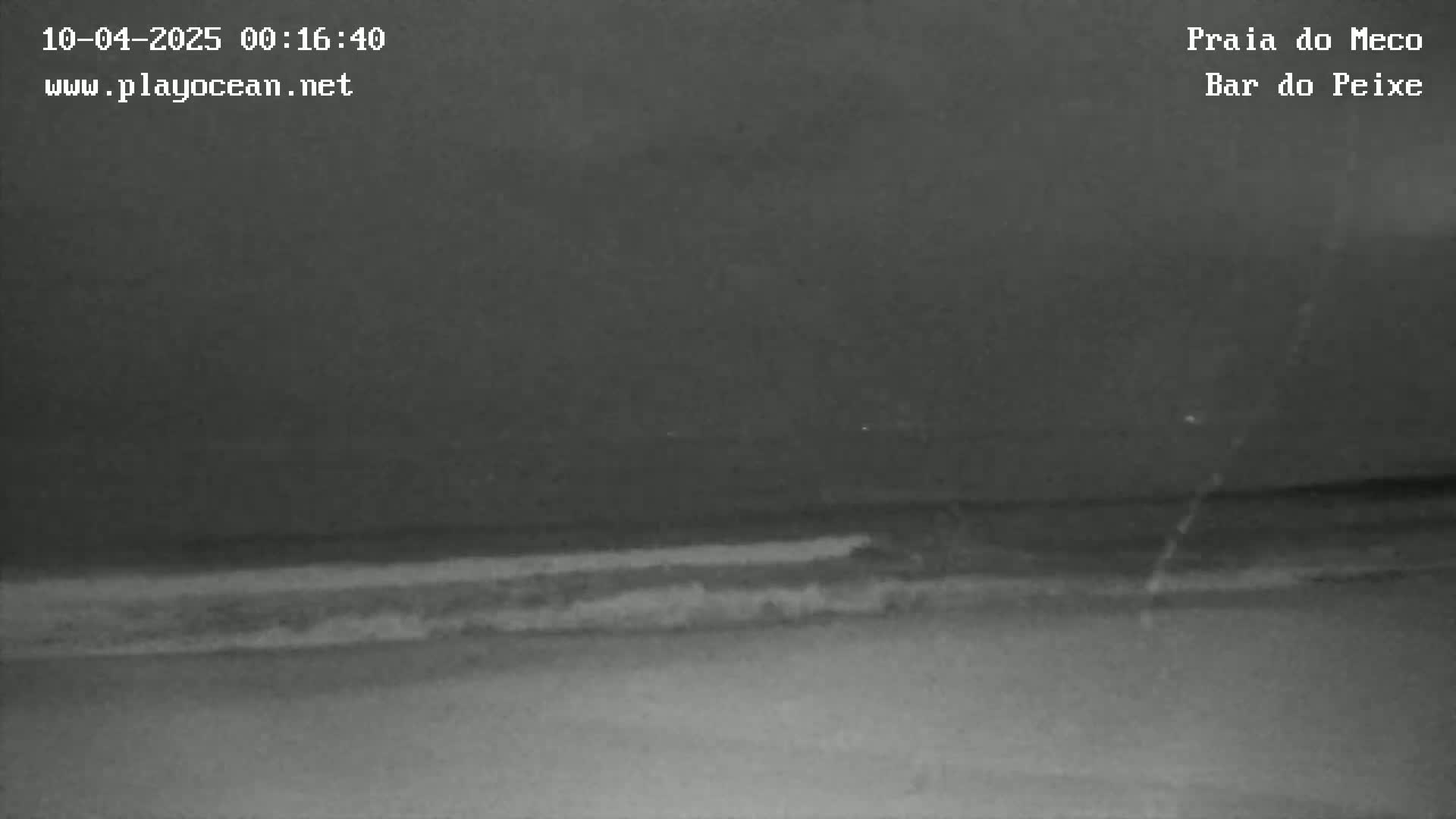 A dark, nighttime beach scene shows gentle waves lapping the shore under a cloudy sky.