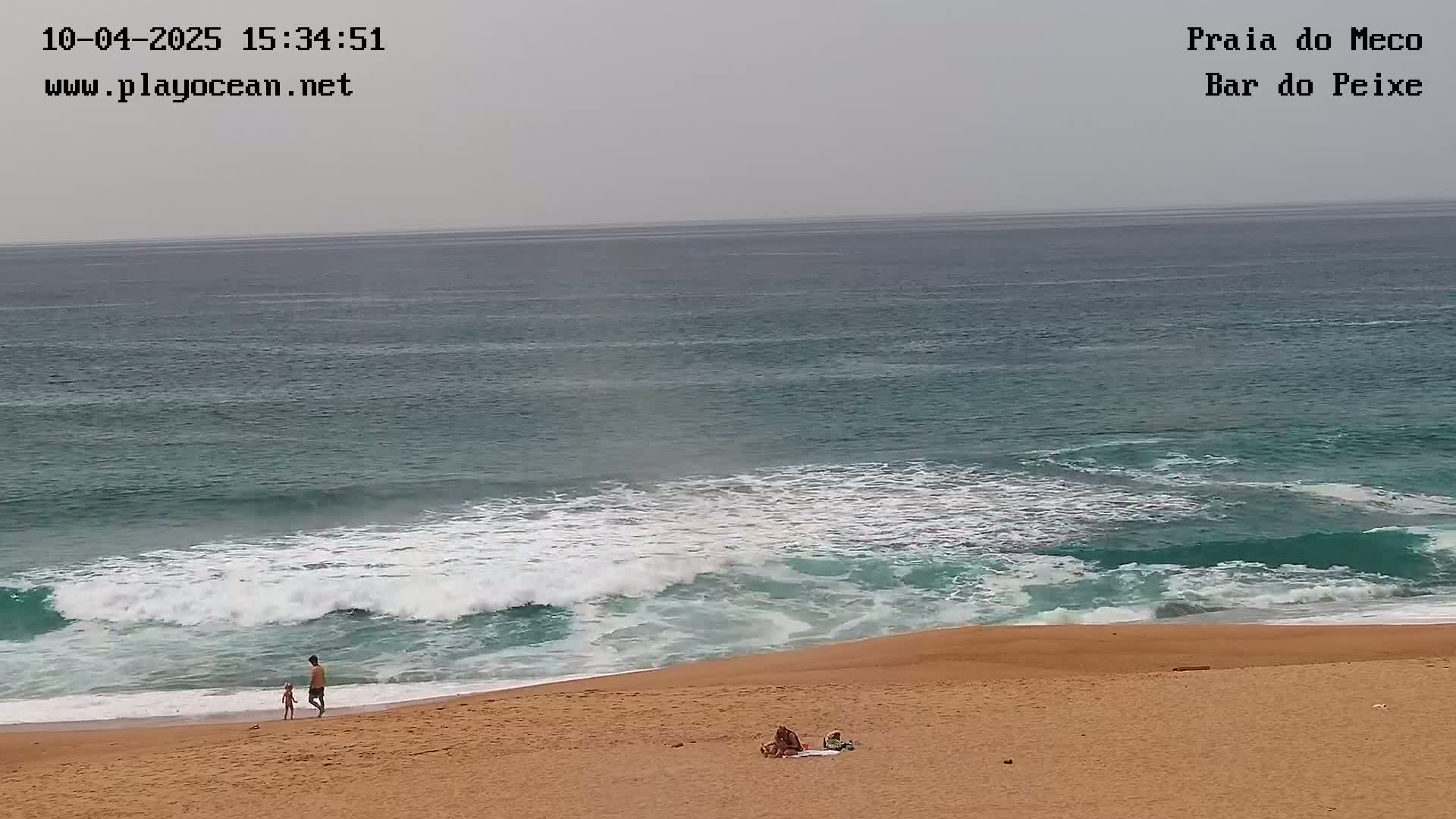 A sandy beach with a few people on it is met by teal-green ocean waves under an overcast sky.
