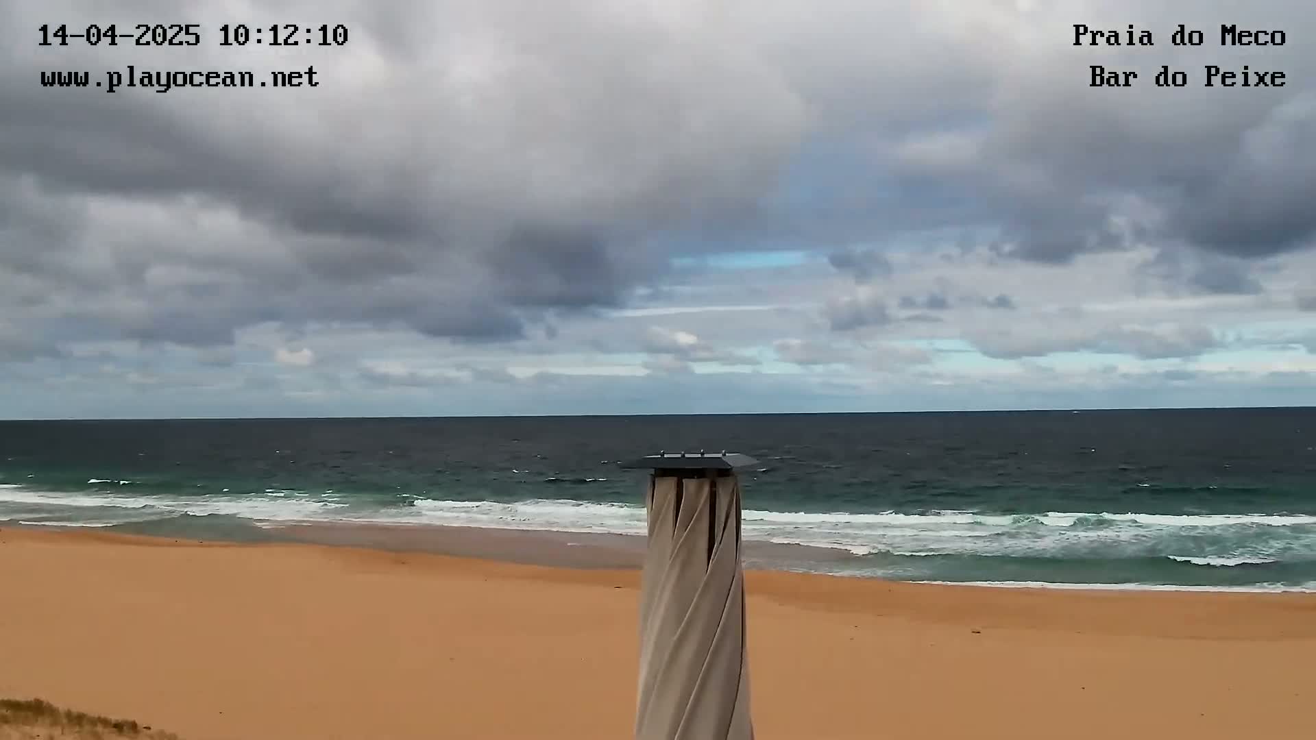 A sandy beach with gentle waves under a mostly cloudy sky is viewed from behind a tall, slender, spiraling beach umbrella.