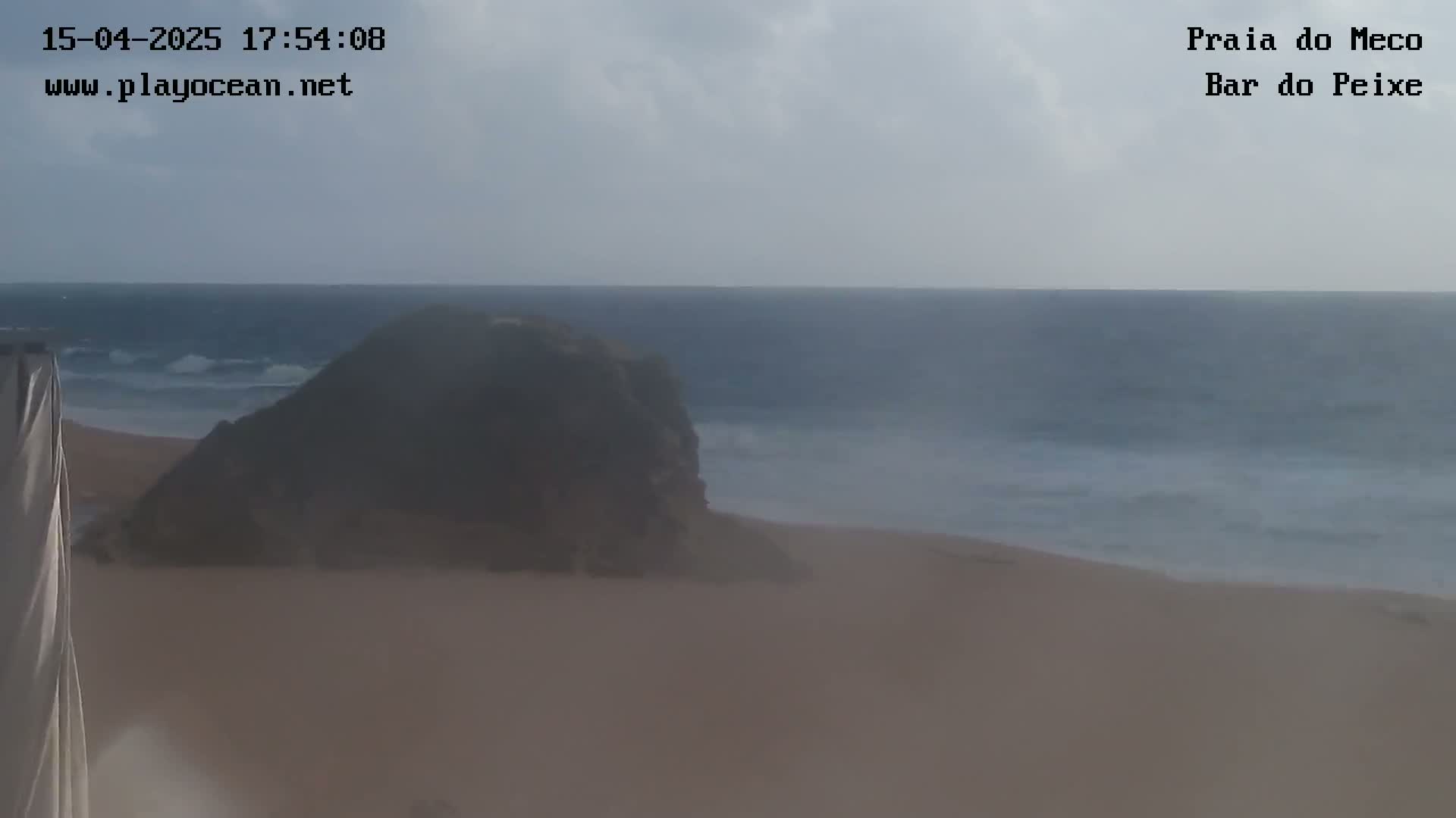 A sandy beach with a dark rock formation is visible, with a somewhat cloudy sky and a calm ocean with small waves in the background.